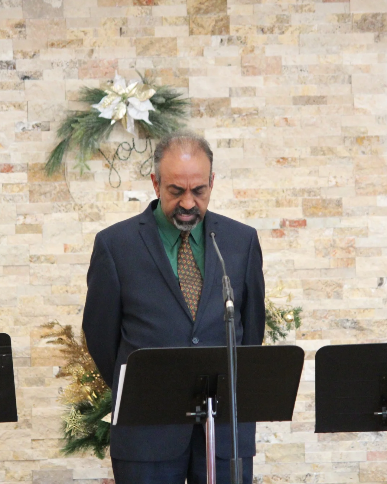 A man in a dark suit and green shirt, with a decorated tie, is standing behind a lectern with a microphone. He is looking down with a serious expression. There is a Christmas wreath and decorations in the background.