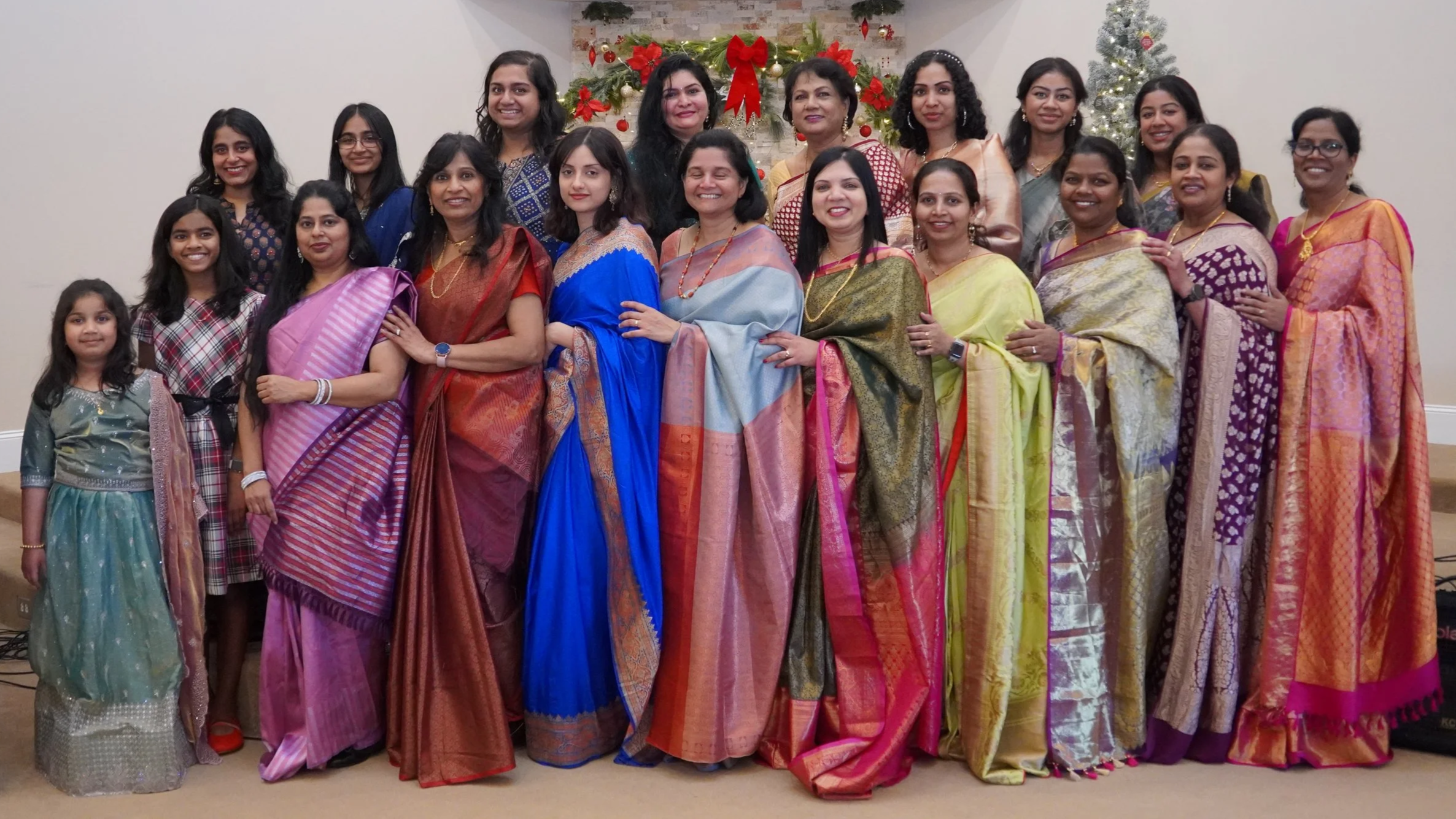 Group of women and children dressed in colorful Indian sarees standing together in front of Christmas decorations, including a decorated tree and red bows.