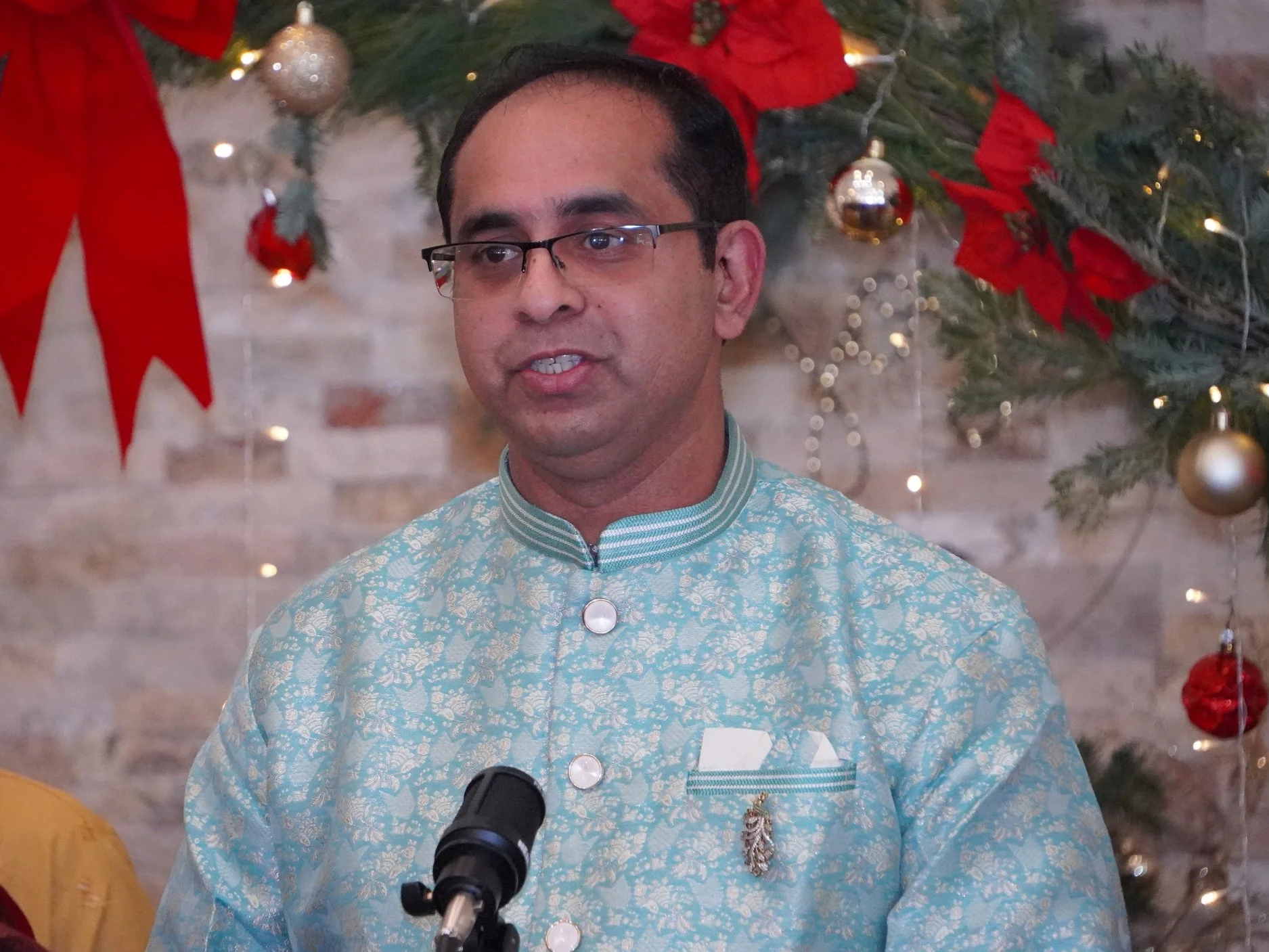 A man wearing glasses and traditional attire, speaking into a microphone with Christmas decorations in the background.