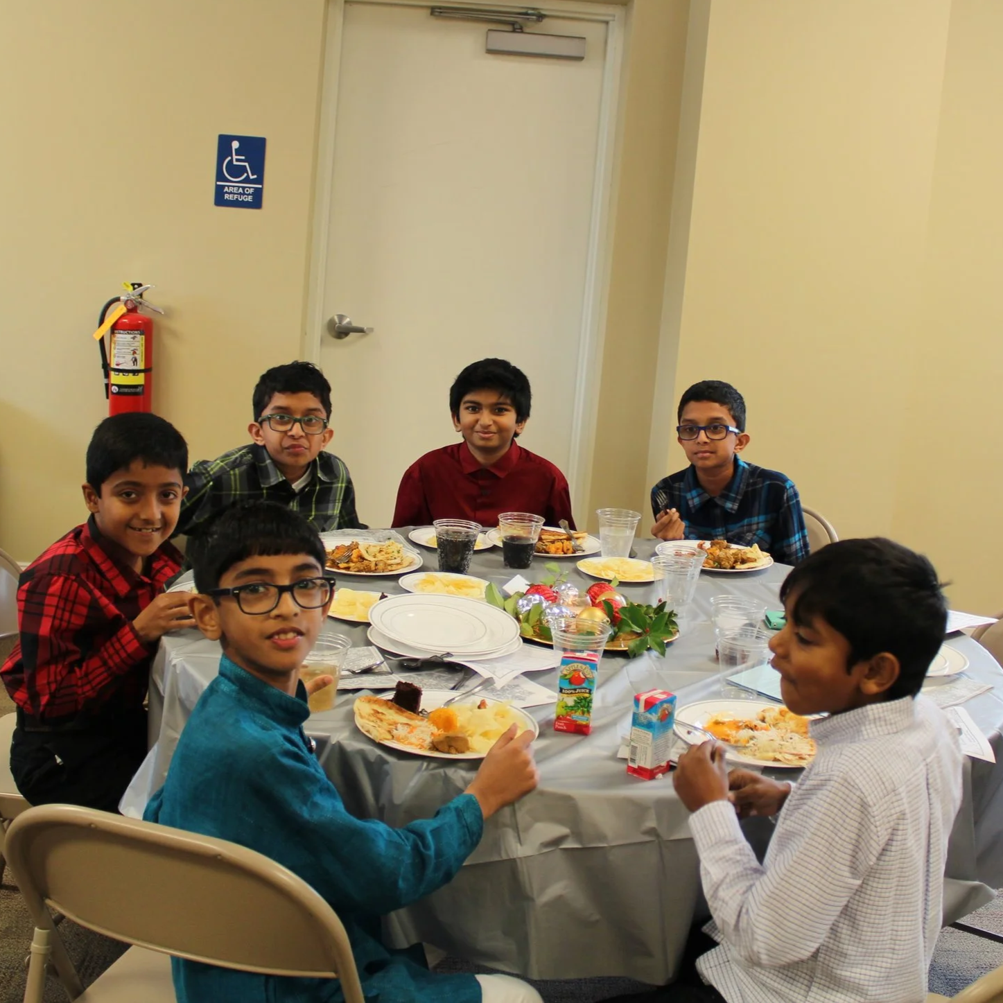 Group of young boys sitting around a table with plates of food, smiling and enjoying a meal at a party or gathering.