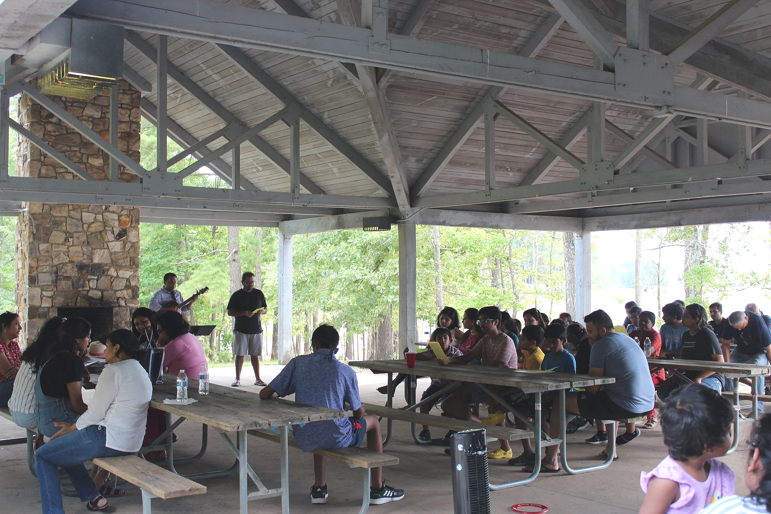 People gathered in a pavilion with a stone fireplace, sitting at picnic tables, attending a community event or gathering, with a person playing guitar and another standing with a microphone, surrounded by trees.