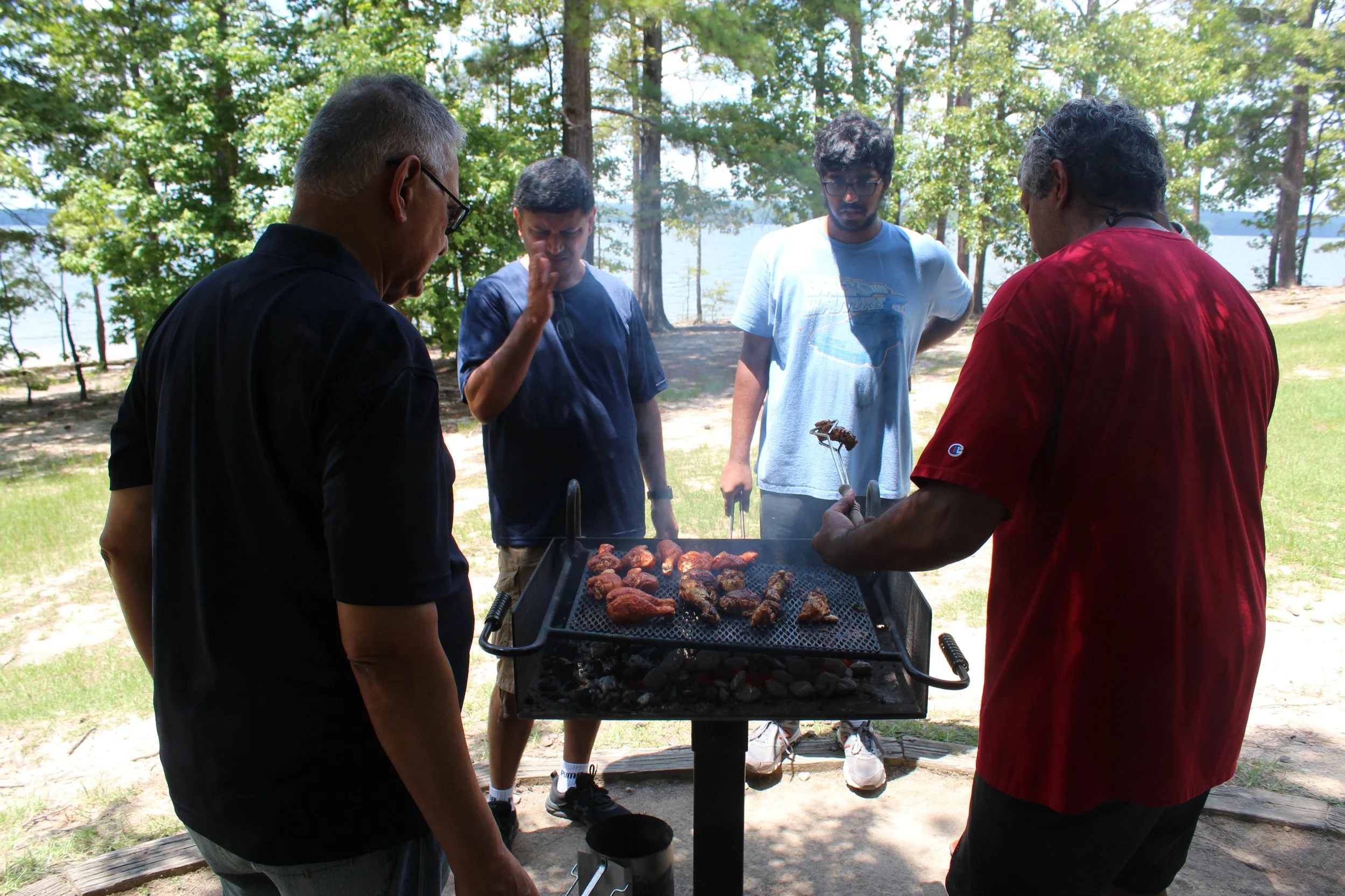 Several people gather around a barbecue grill outdoors, cooking meat, with trees and a lake in the background.