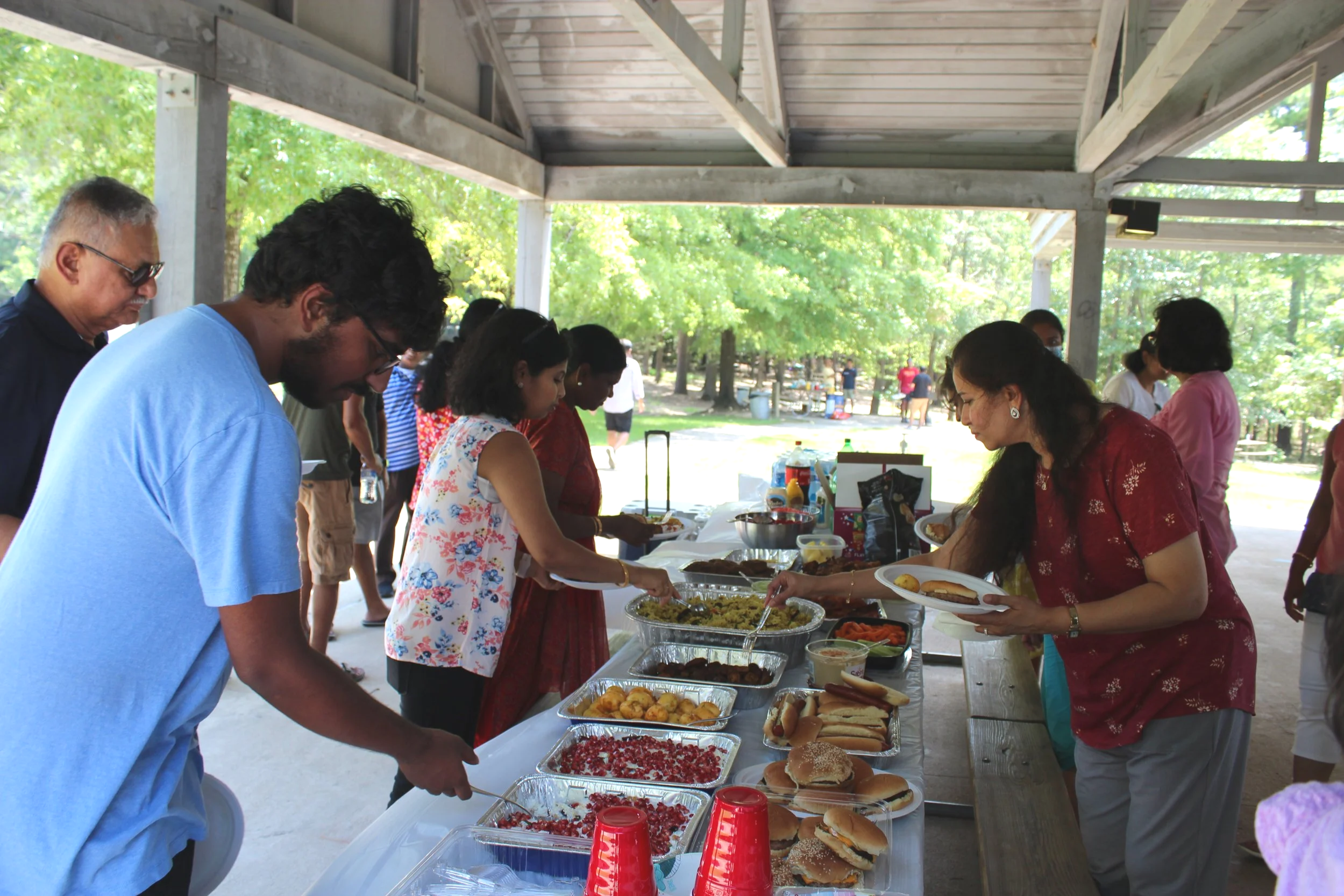 People at a picnic buffet table outdoors under a pavilion, serving themselves food such as hot dogs, salads, and desserts.
