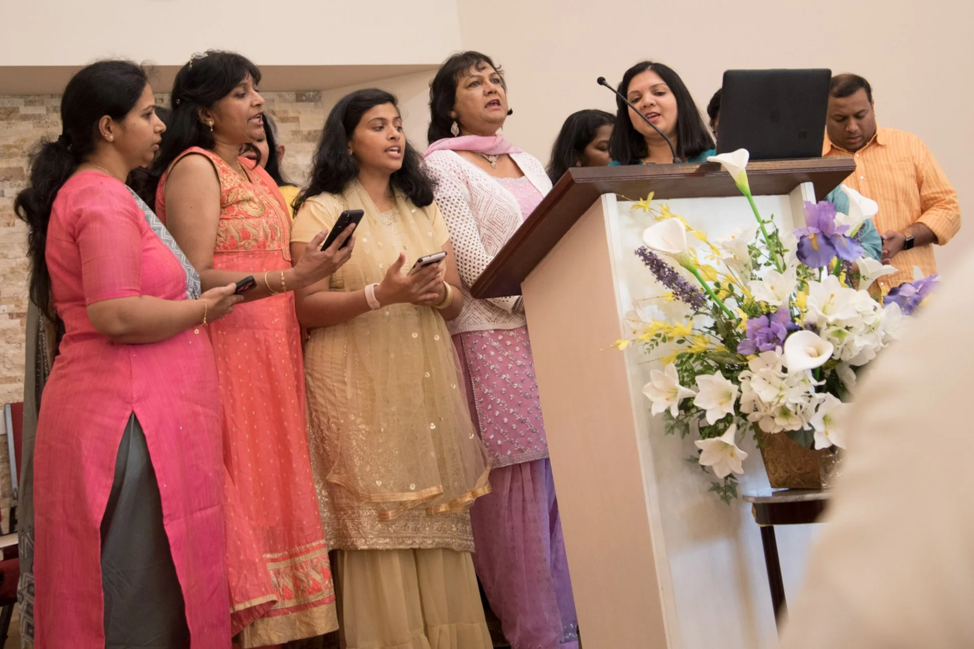 Group of women dressed in traditional attire, singing at a church or community event with a floral arrangement in front of a podium.