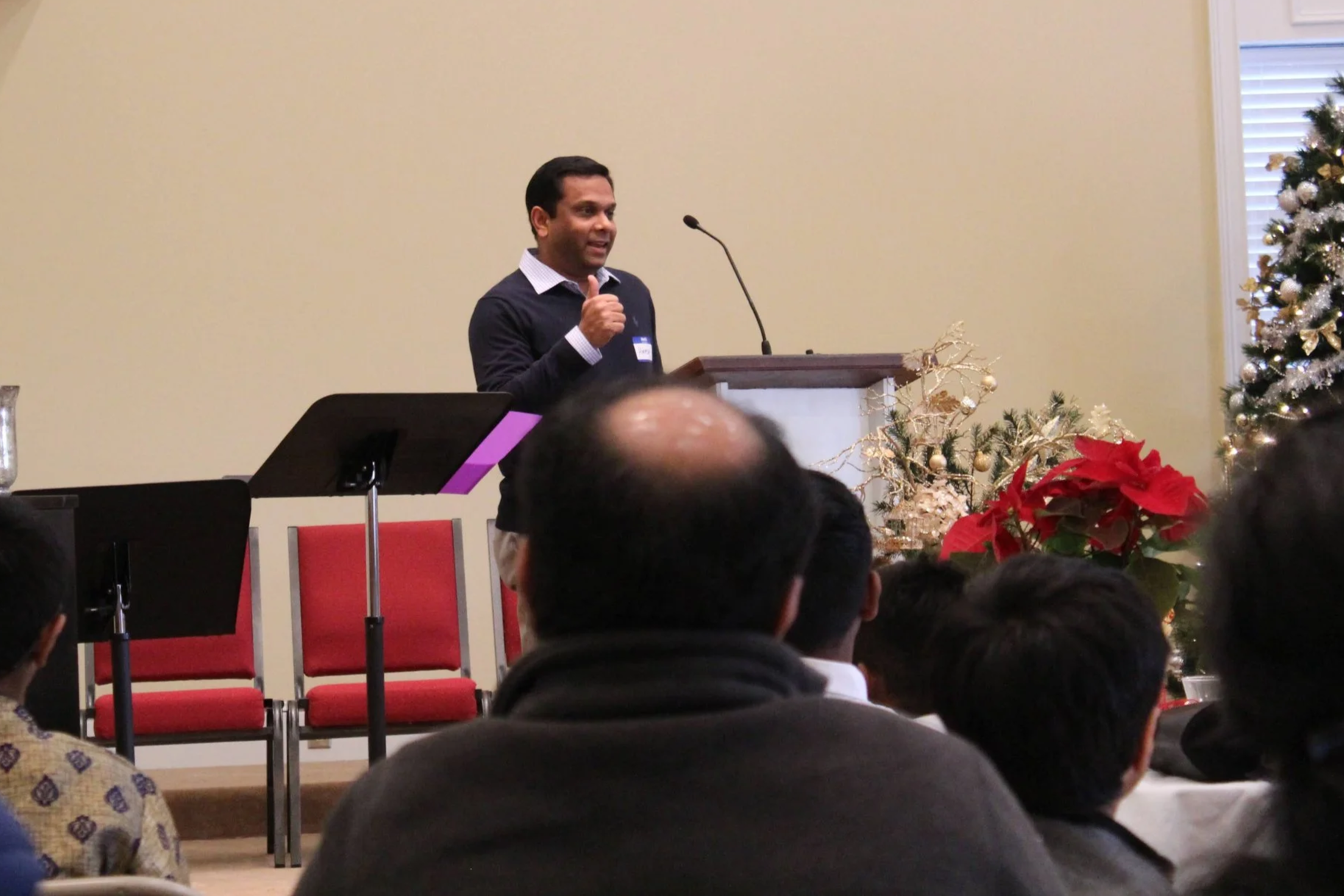 Man speaking at a podium during a holiday event, decorated with Christmas trees and poinsettias.