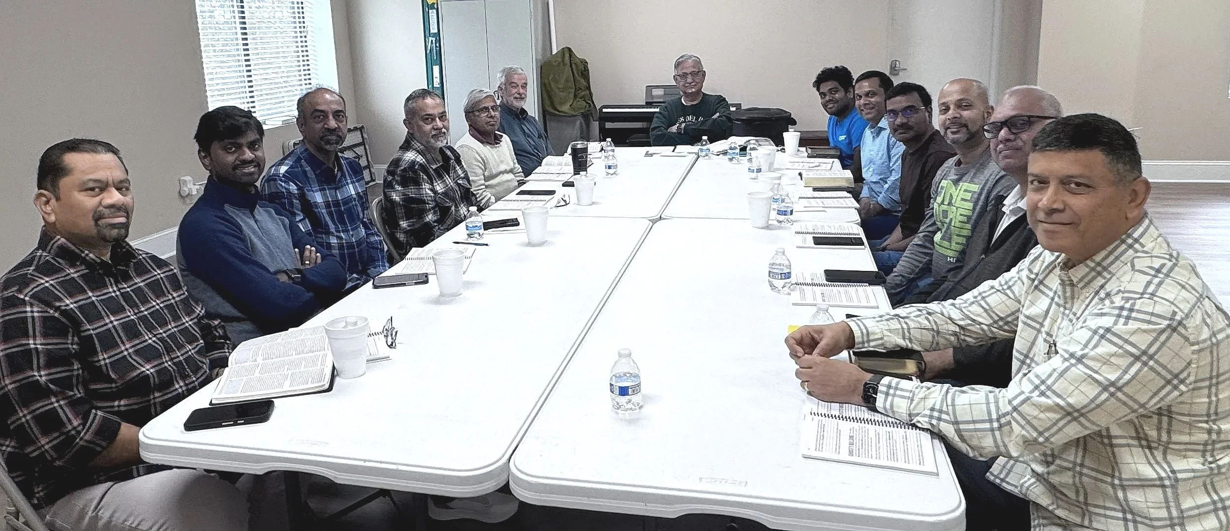A group of fifteen men sitting around a conference table in a meeting room, with documents, water bottles, and cups on the table.