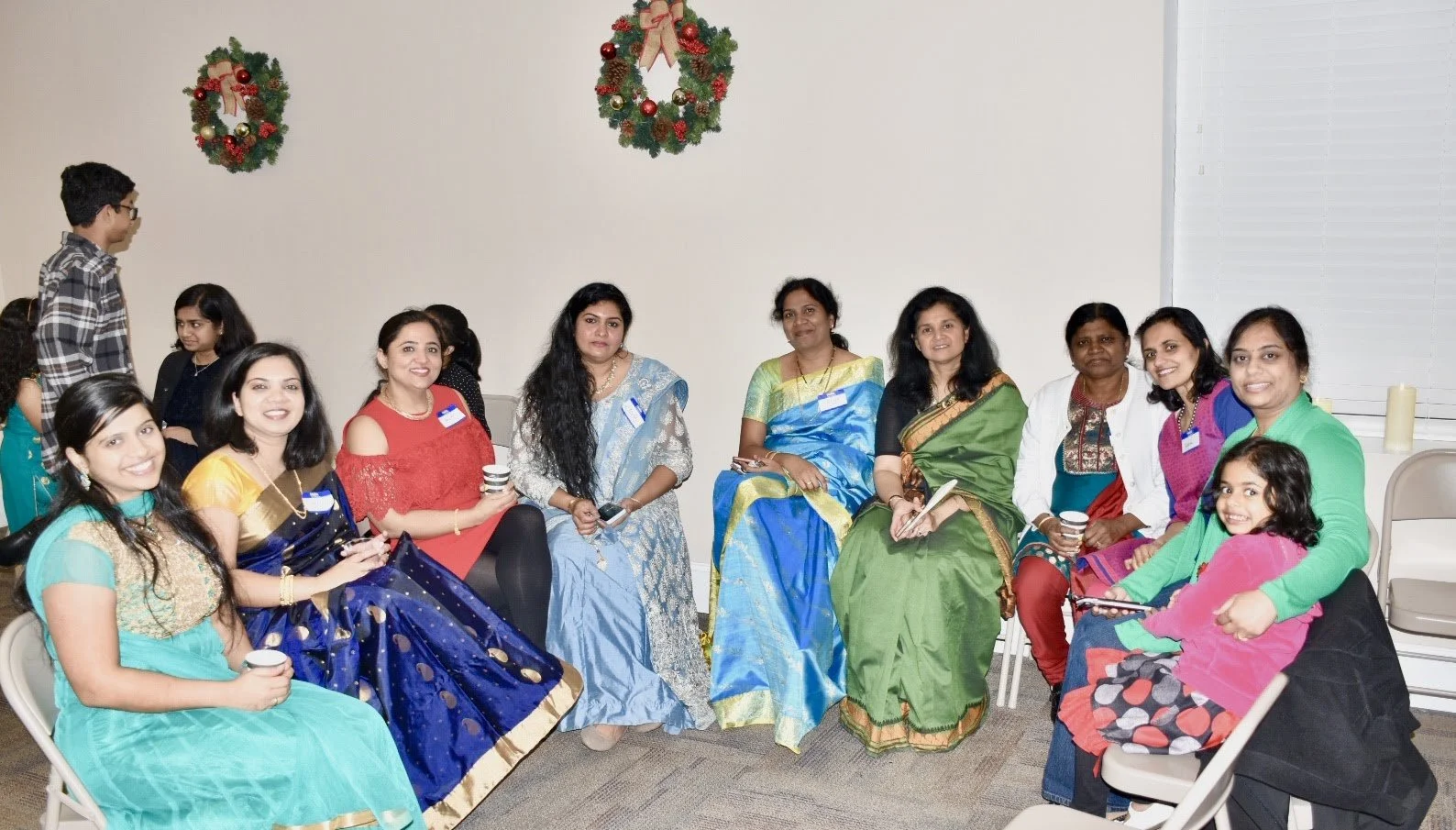 Group of women and children gathered indoors during a festive event with Christmas wreaths on the wall.