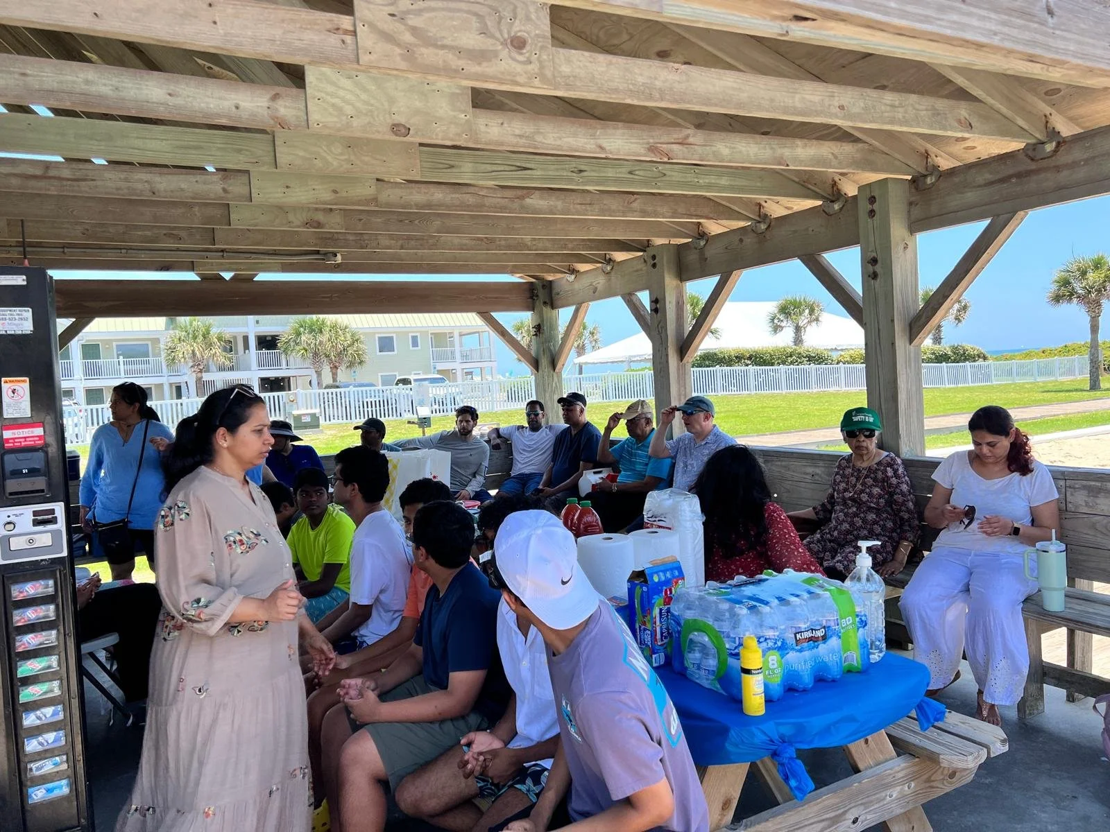 Group of people gathered under a wooden pavilion at a beachside location, sitting on benches and engaging in conversation, with bottled water and supplies on a table, palm trees and beach in the background.