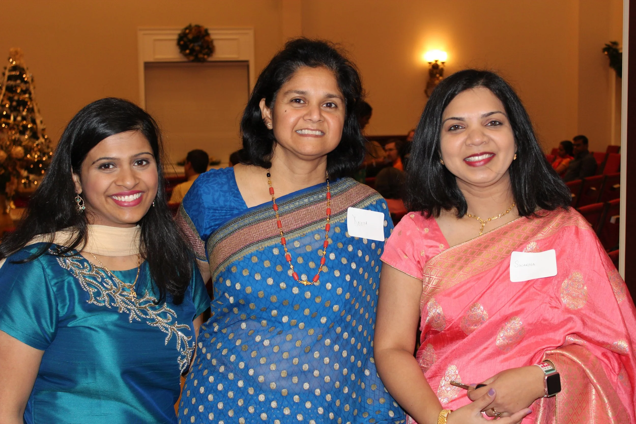 Three women of Indian ethnicity smiling at an indoor event, dressed in colorful sarees, with festive decorations including a Christmas tree in the background.