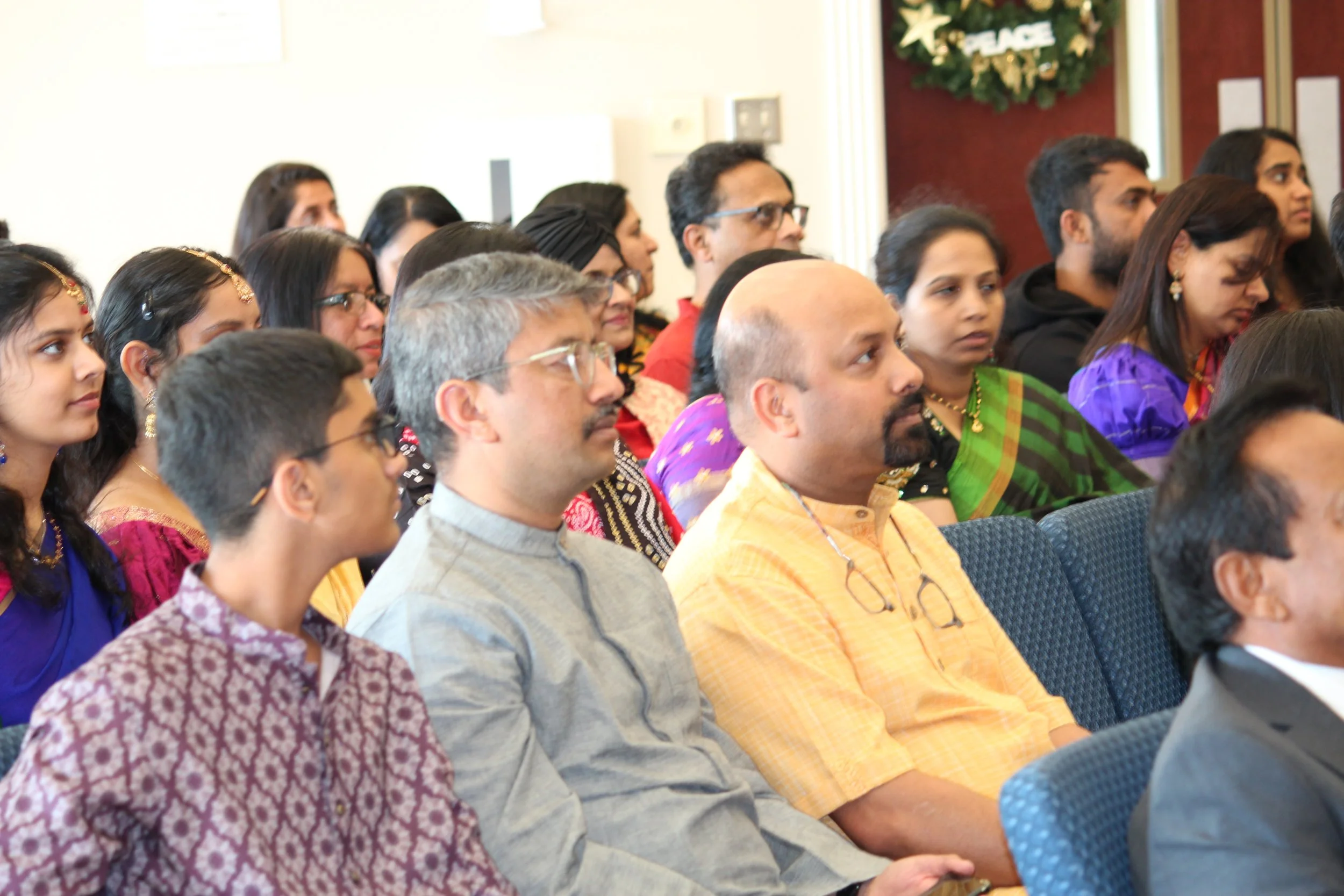 Audience attending an indoor event, seated and listening attentively, diverse group including men and women of various ages and ethnicities, some wearing traditional Indian attire, in a decorated room with a Christmas wreath that says 'PEACE' in the 