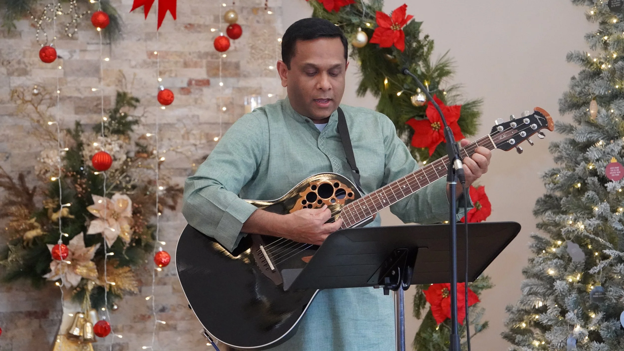 A man playing a black and gold acoustic guitar at a Christmas-themed setting with decorated trees and wreaths.