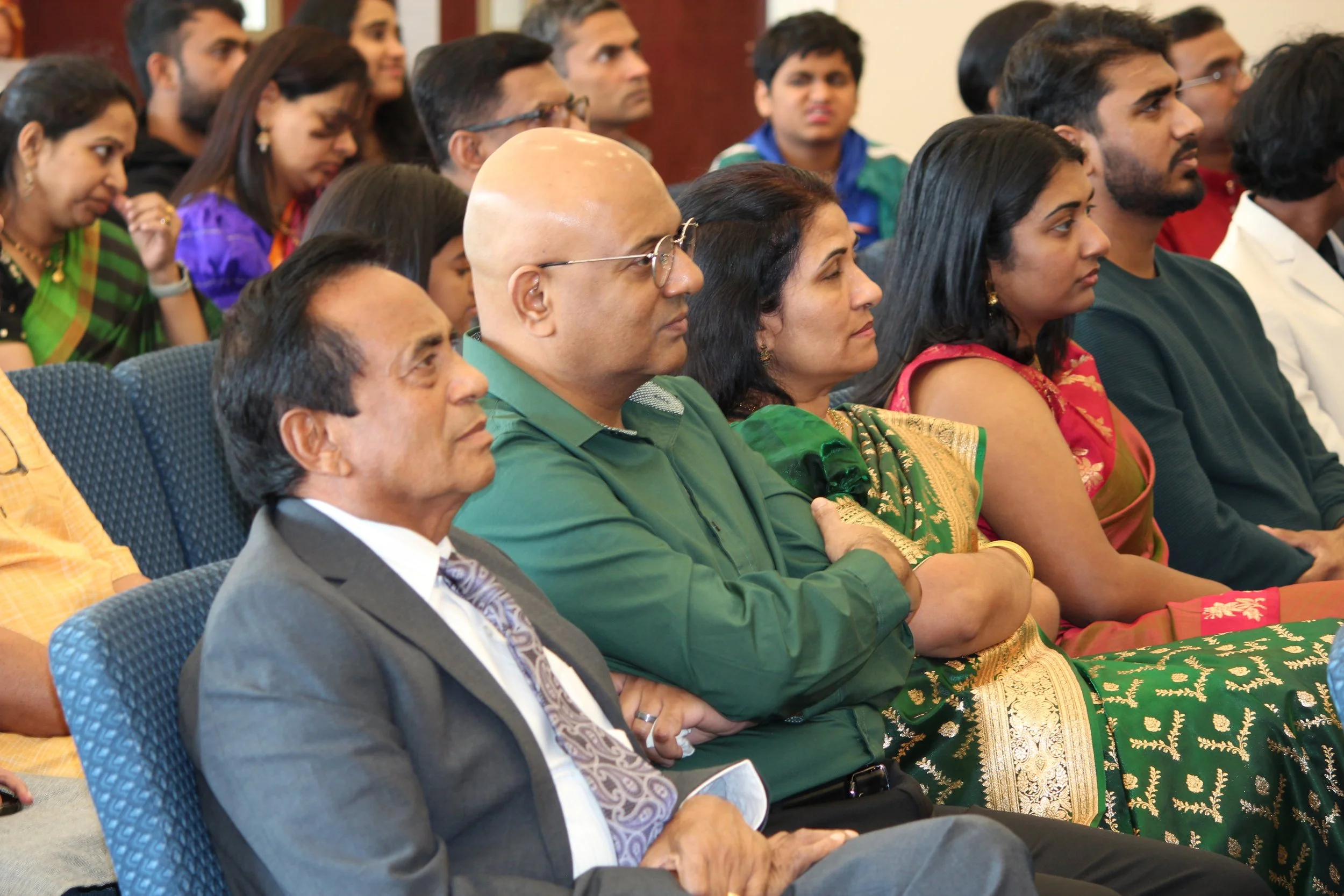 Audience of men and women sitting attentively during a formal event. Some are dressed in traditional Indian attire, others in business suits.
