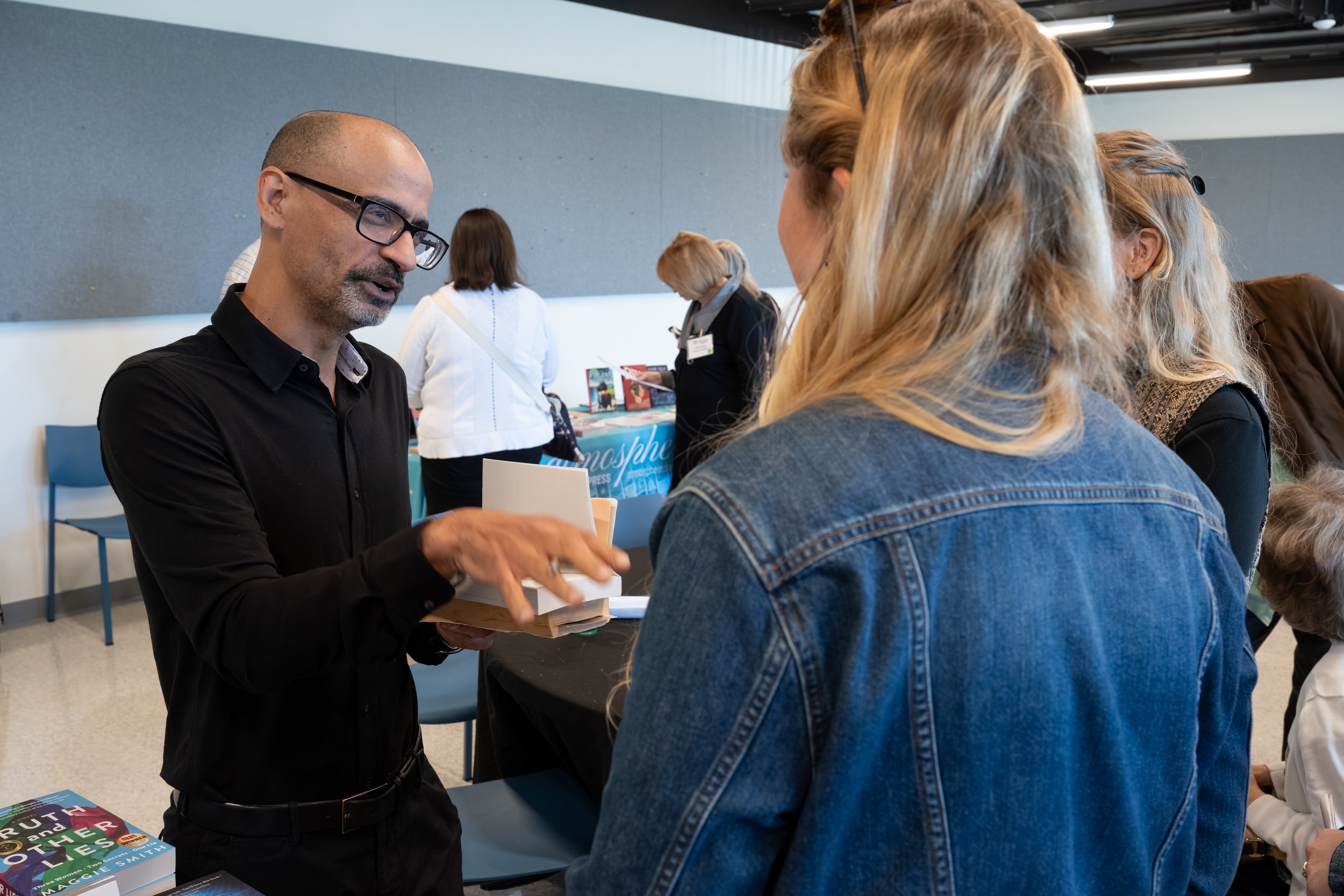 A man with glasses signing a book for a woman with long blonde hair wearing a denim jacket at a book event or signing.