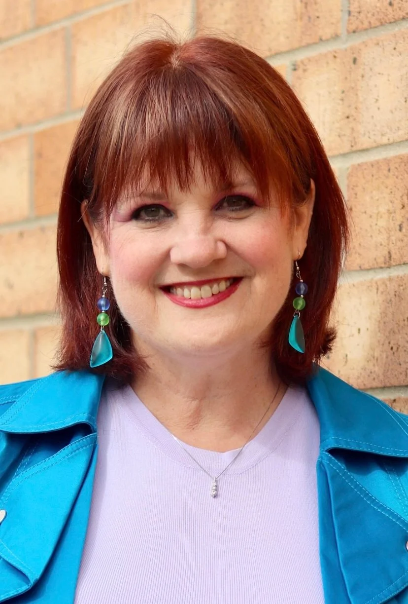 A woman with red hair and earrings, standing outdoors in front of a brick wall, smiling at the camera.