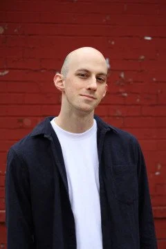 A bald man in a dark jacket and white shirt stands against a red wooden wall, looking at the camera with a slight smile.