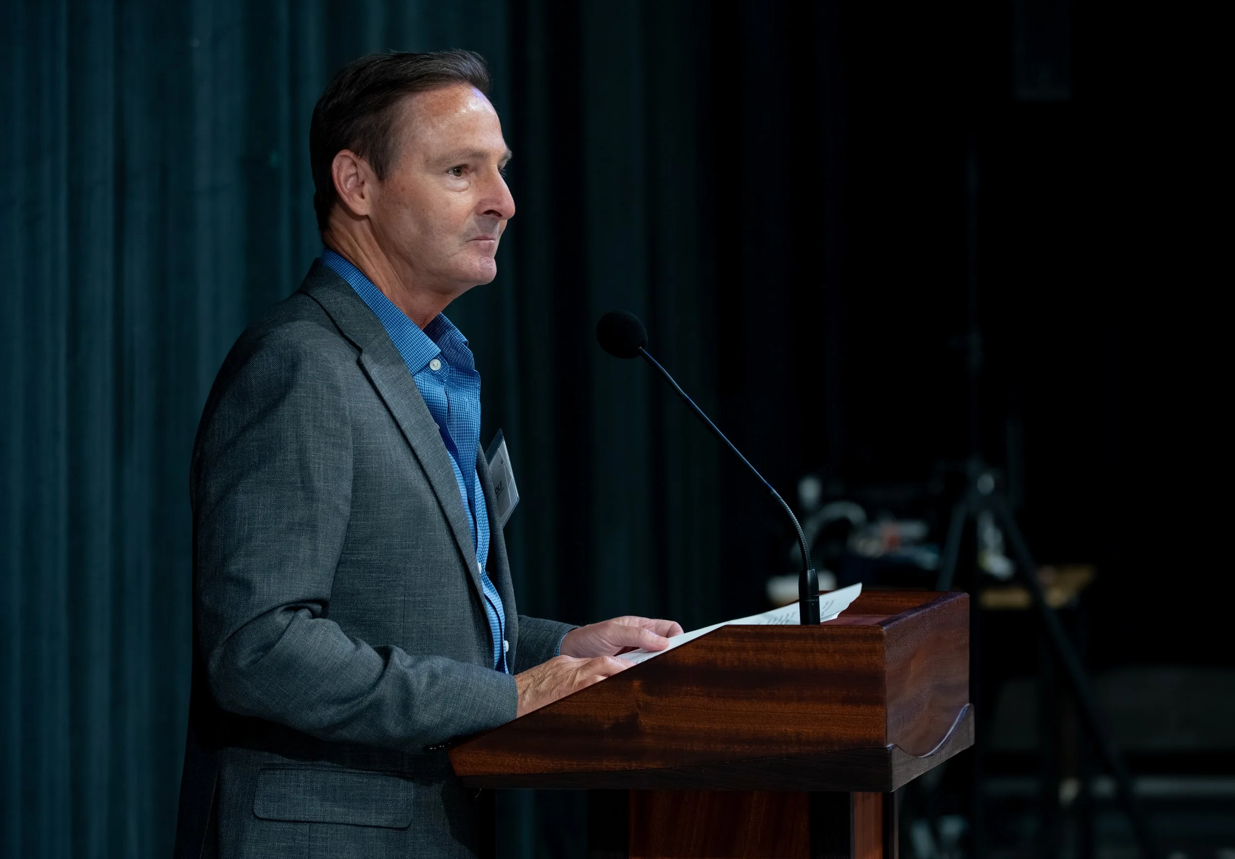A man in a gray suit and blue shirt speaking at a podium with a microphone.