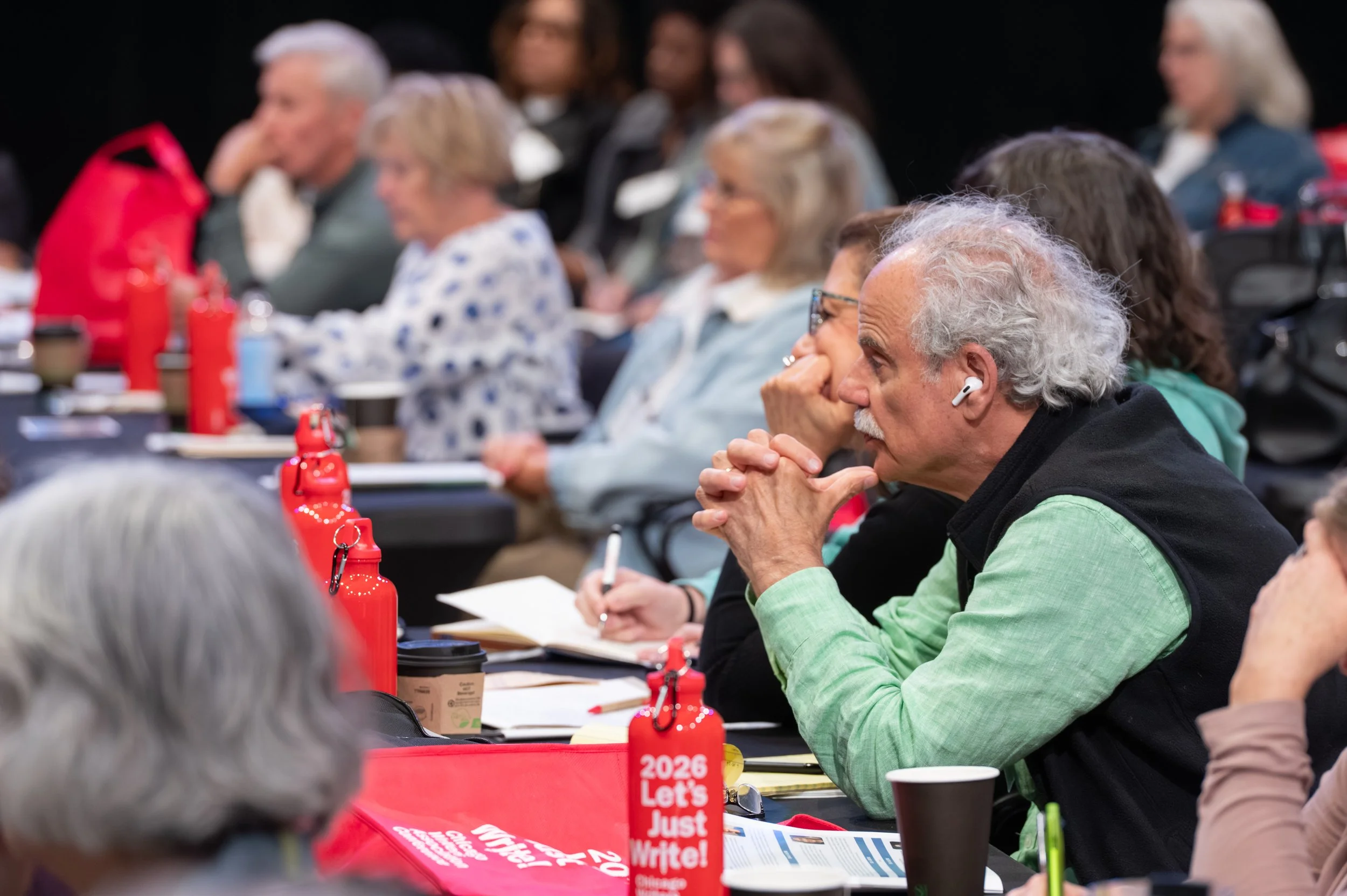 A diverse group of people sitting at a long table during a meeting or conference, attentively listening and taking notes, with red water bottles and coffee cups on the table.