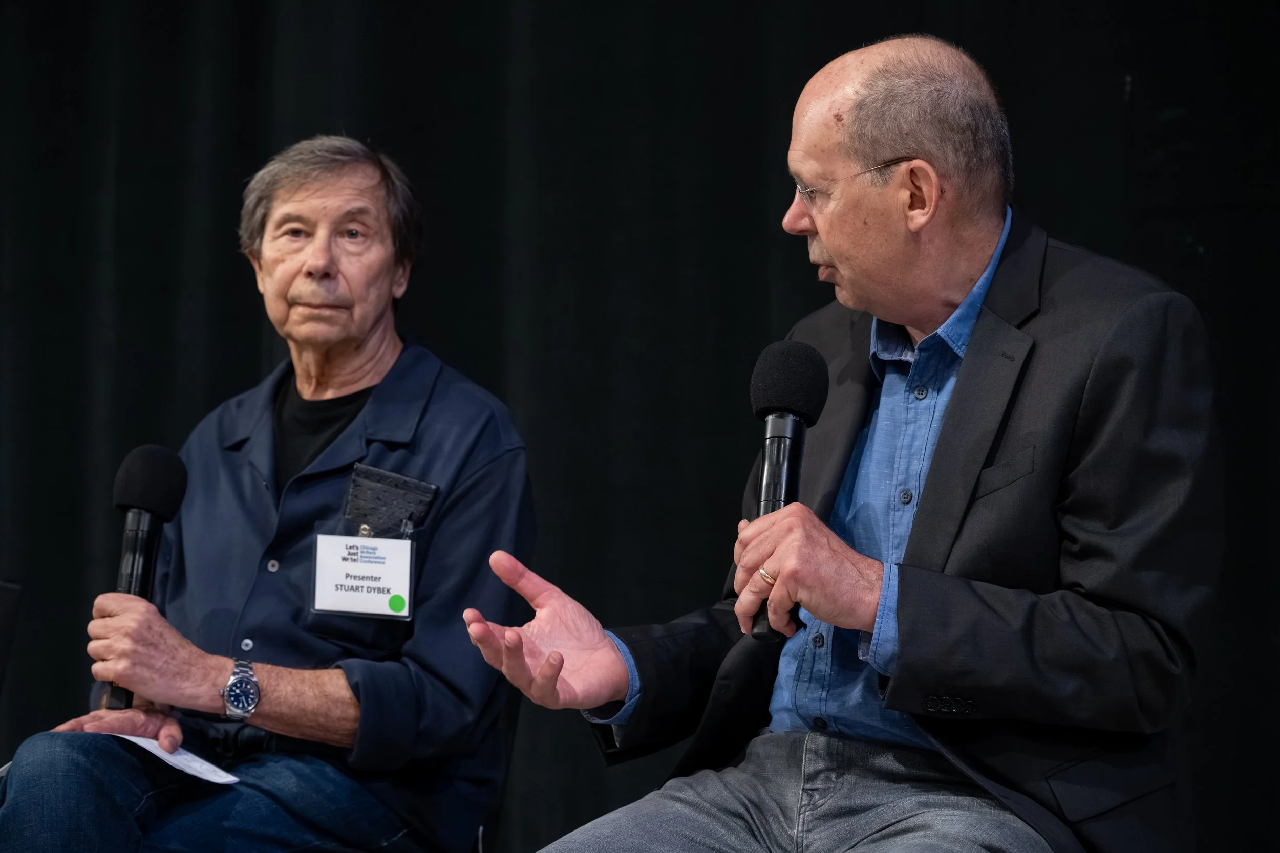 Two men are participating in a panel discussion or interview, each holding a microphone. The man on the left is older with gray hair, wearing a navy blue shirt with a name badge, and listening attentively. The man on the right is bald, wearing glasse