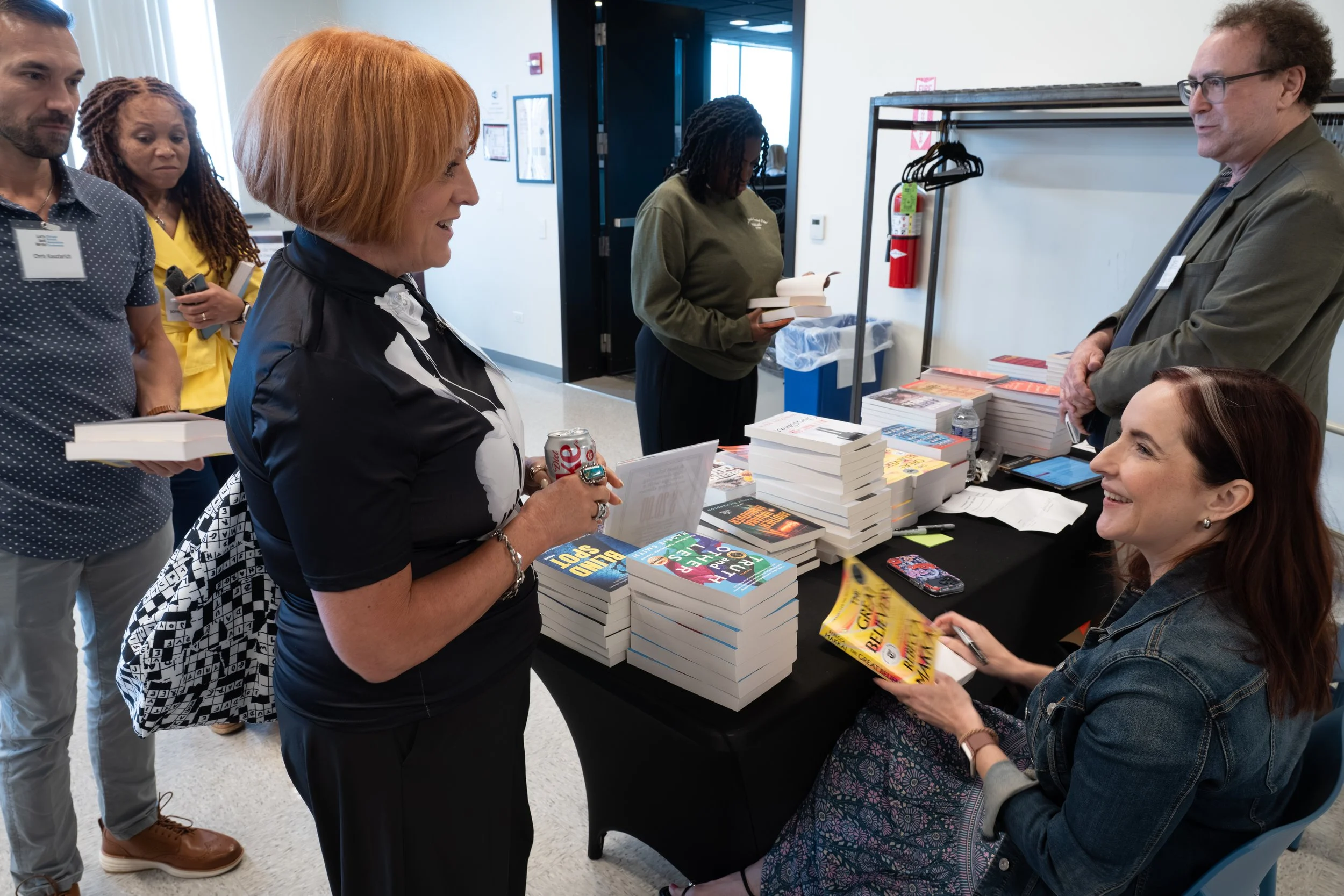 A group of people at a book signing event, with an author signing books and engaging with attendees. Books are displayed on a table.