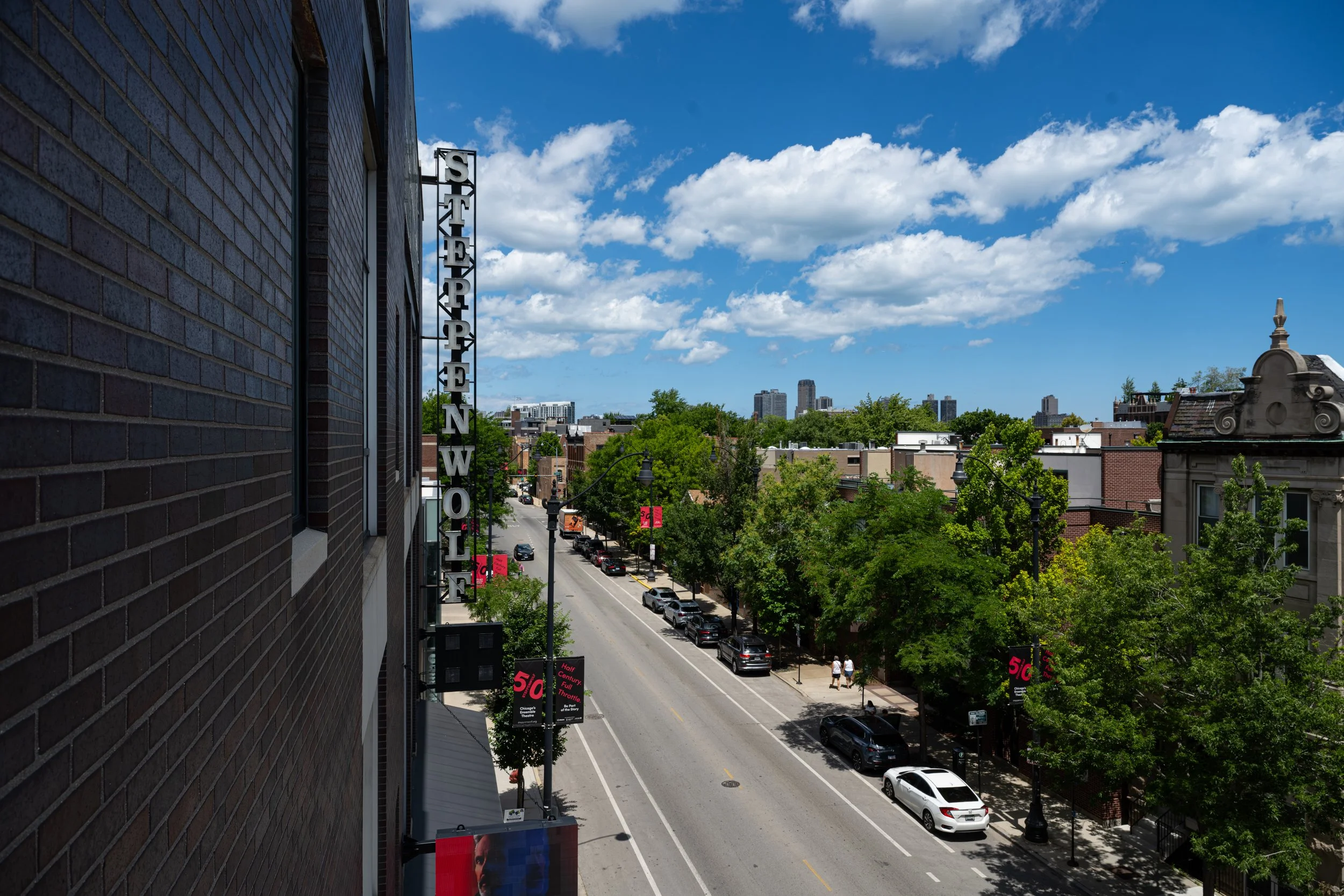 View of a city street with parked cars, trees, and buildings under a partly cloudy sky, seen from an upper floor of a building with dark brick exterior and vertical signage reading 'STUDIO W'.