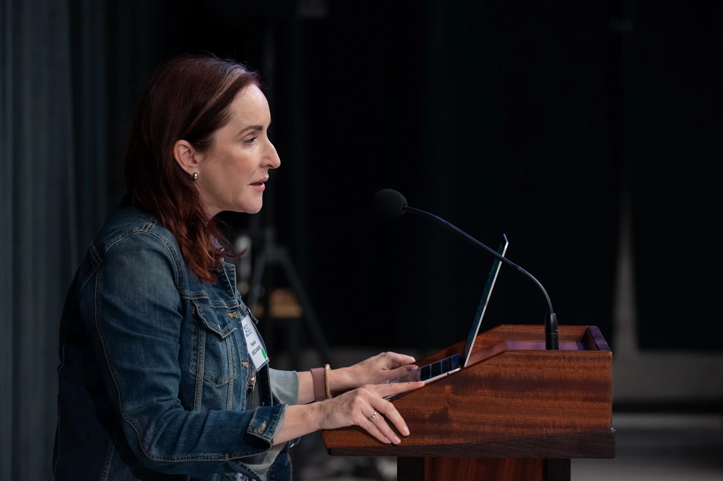 A woman with long reddish-brown hair, earrings, and a denim jacket is speaking at a wooden podium with a microphone in a dark room with a black background.