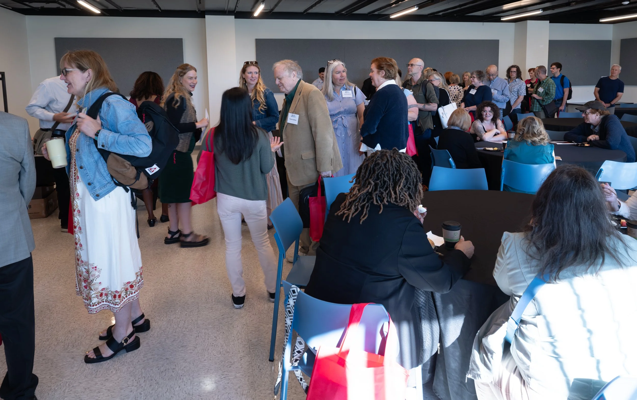 People gathered in a conference room, some standing and chatting, others seated at tables, engaging in discussions.