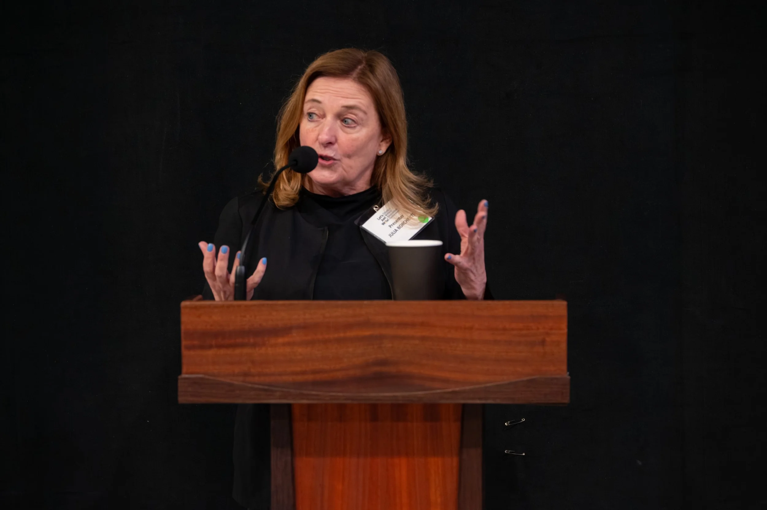 A middle-aged woman with shoulder-length brown hair speaking at a podium, wearing a black top and a name tag, with a black background.