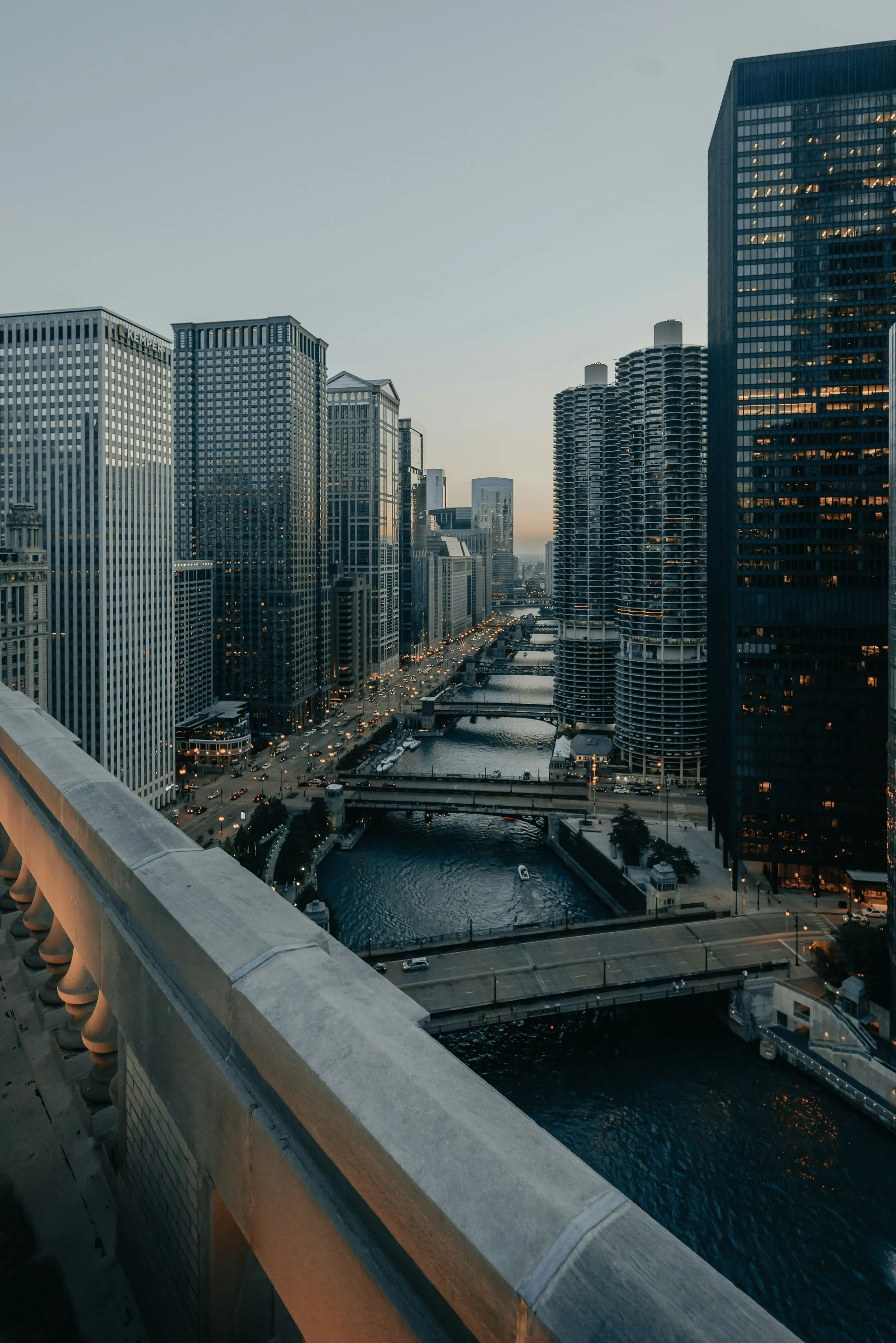View of Chicago downtown skyline at dusk with tall modern buildings, a river with boats, bridges, and city lights starting to turn on.