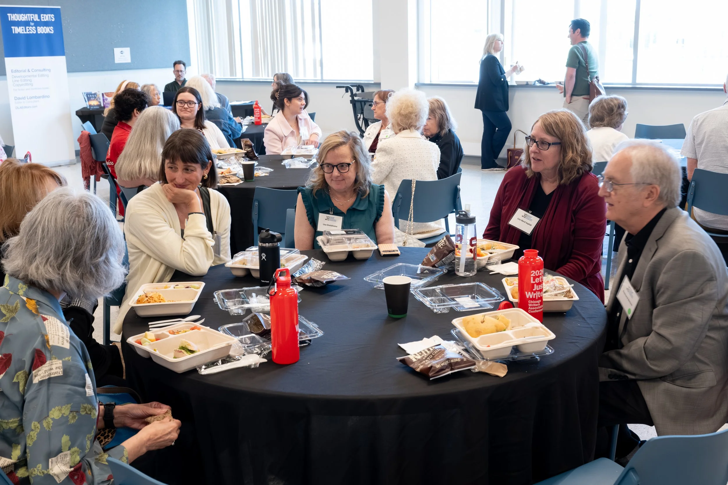 A diverse group of people attending a meal at round tables during a conference or event in a bright room with large windows, with food containers, drinks, and utensils on the tables.