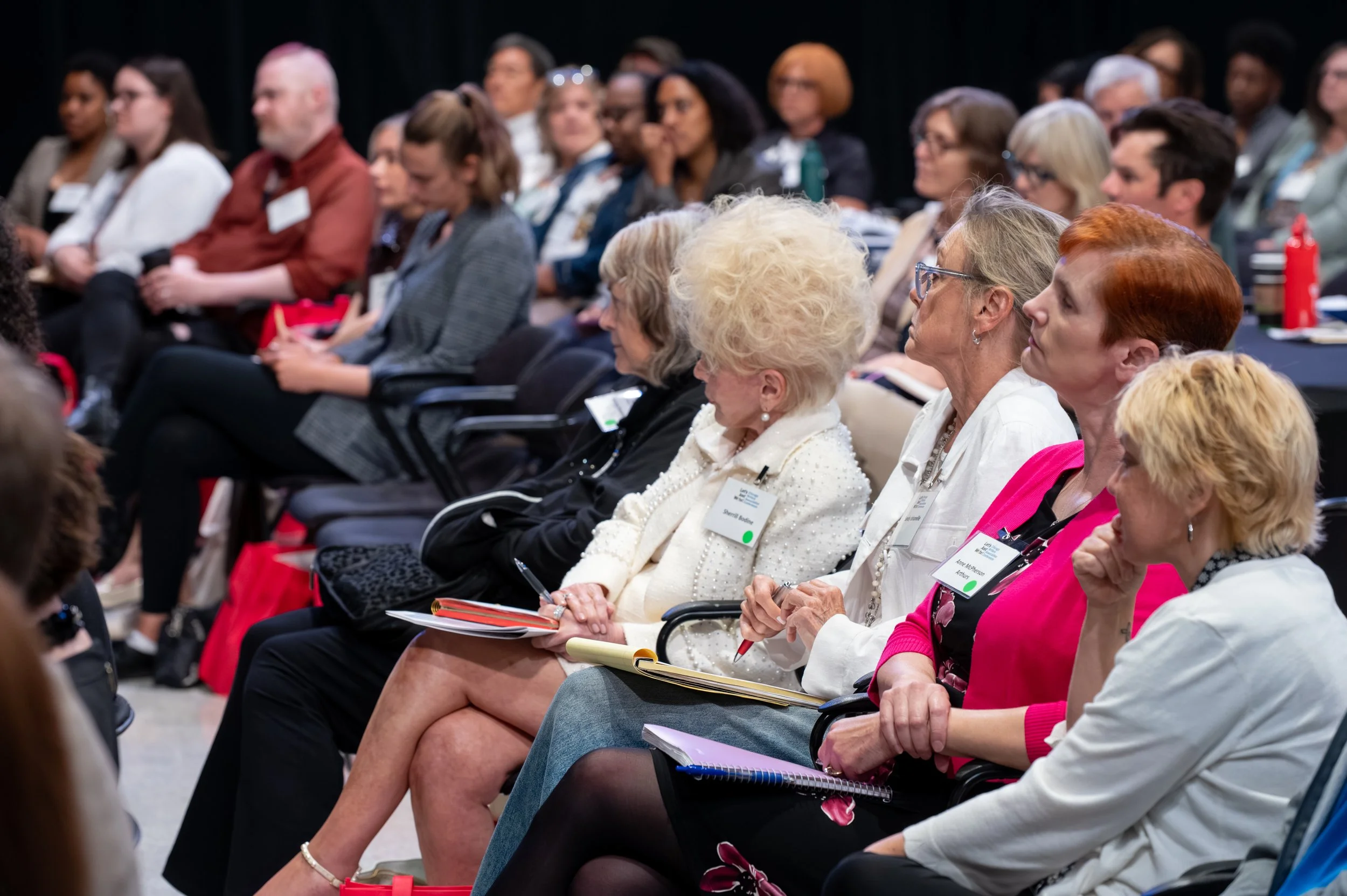 Audience seated in conference room listening to a presentation, with some taking notes and wearing name tags.