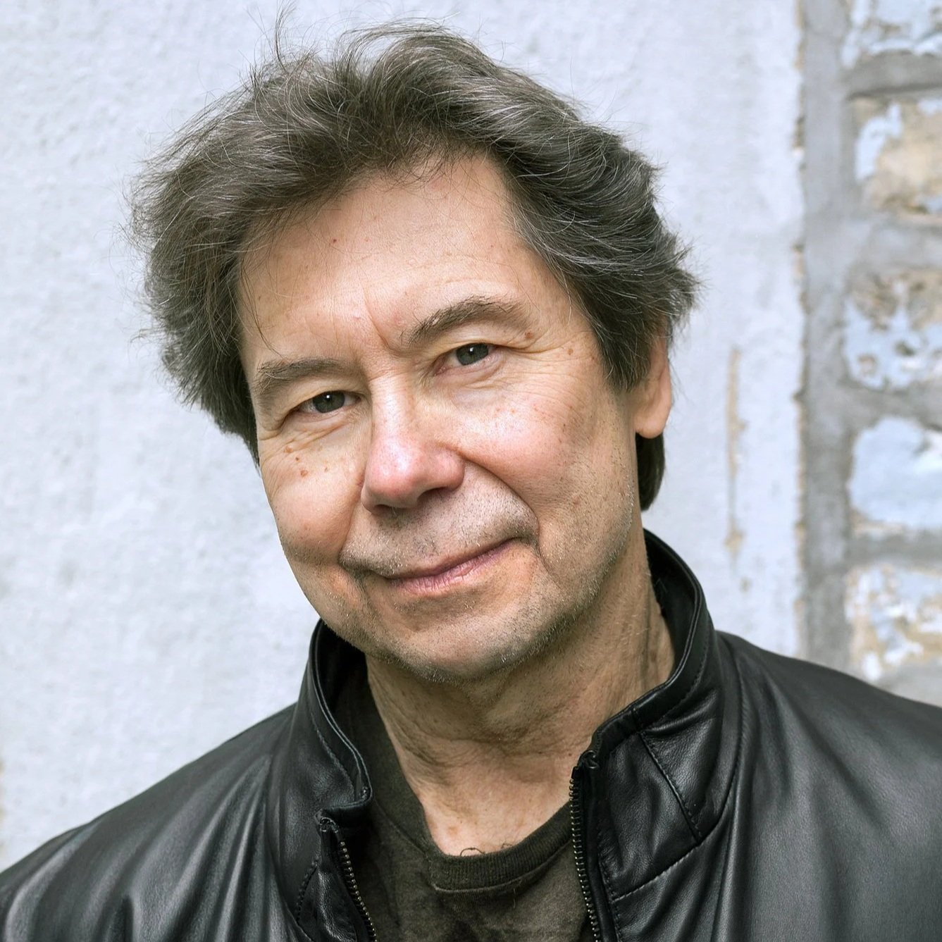 A middle-aged man with dark, slightly wavy hair and blue eyes, smiling gently, wearing a black leather jacket, standing outside against a gray stone wall.