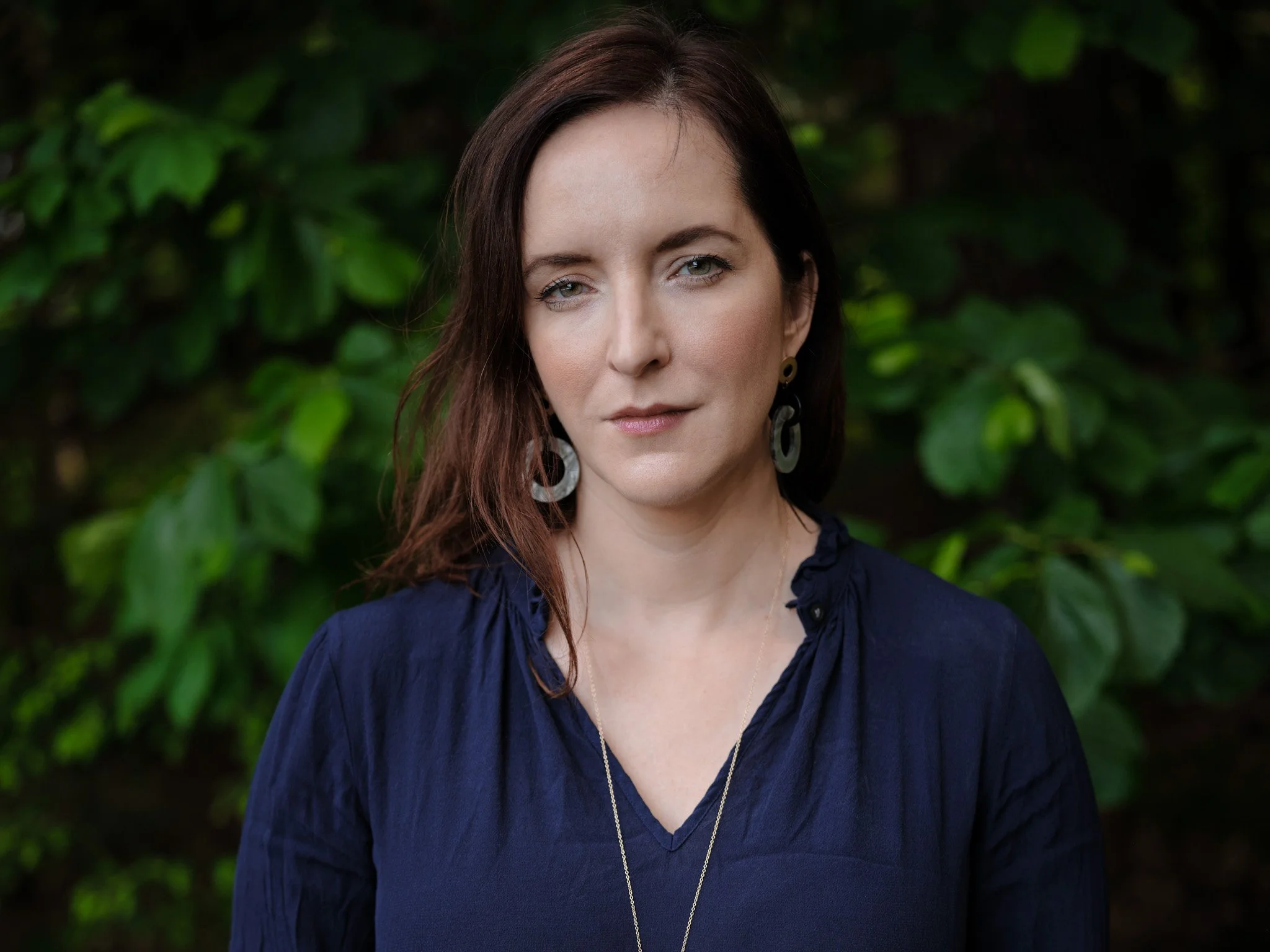 A woman with long, dark red hair, wearing large hoop earrings, a navy blue top with a deep neckline, and a delicate gold necklace, standing outdoors against a background of green leaves.