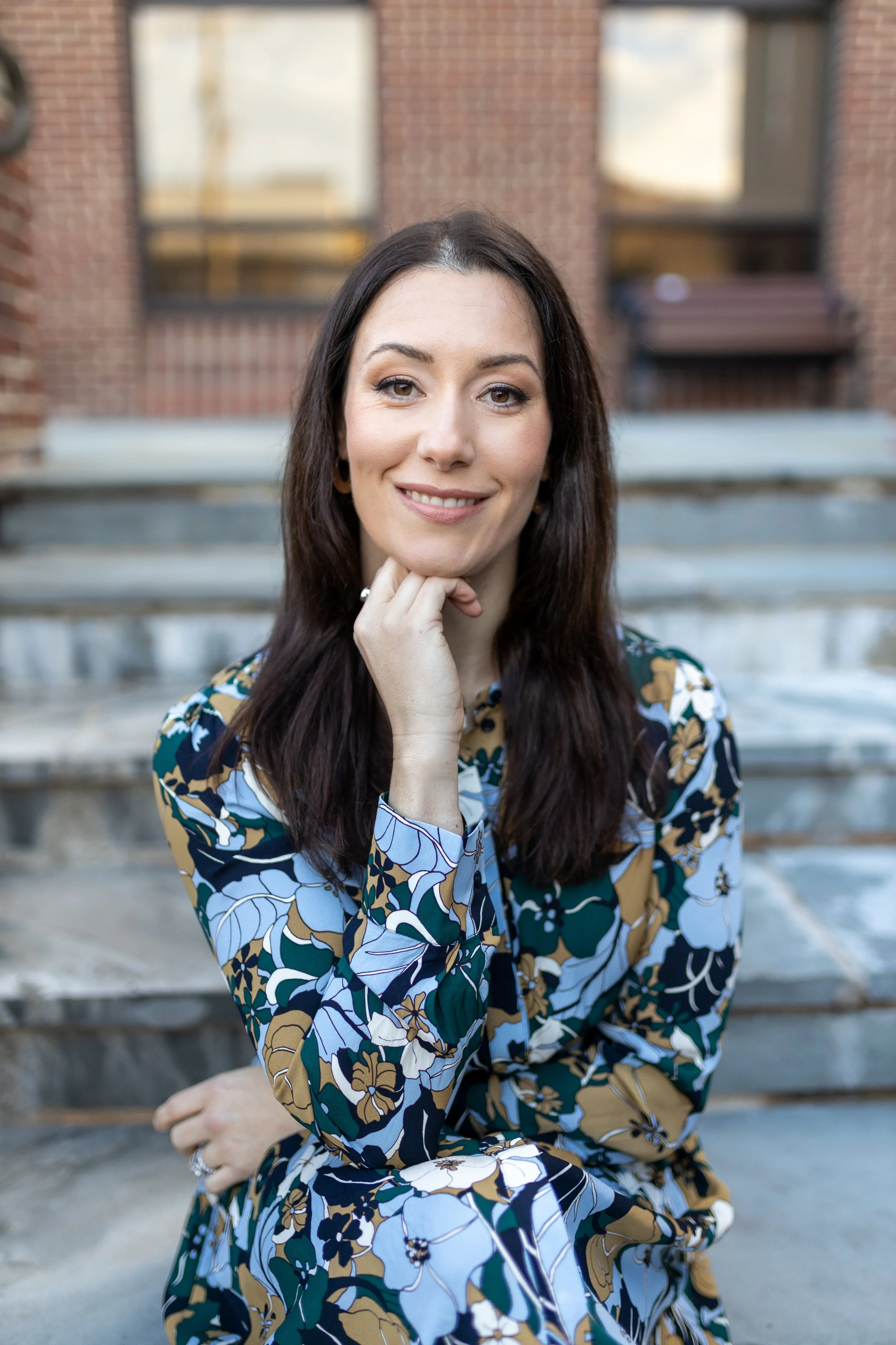 A woman with long dark hair, smiling gently, with her hand resting on her chin, sitting on outdoor steps with a brick building in the background, wearing a colorful floral patterned dress.