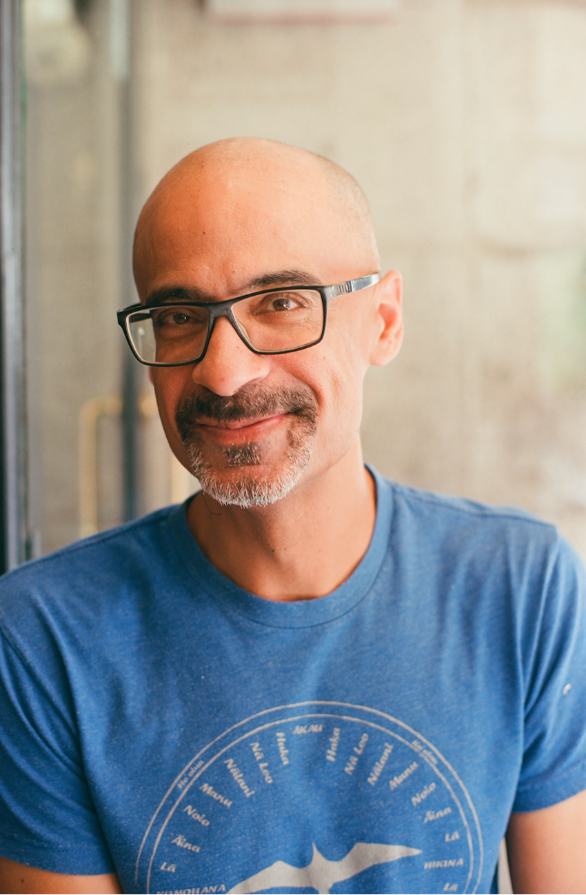 A smiling man with a bald head, glasses, and a light beard, wearing a blue t-shirt with white text and graphics, sitting indoors near a window.