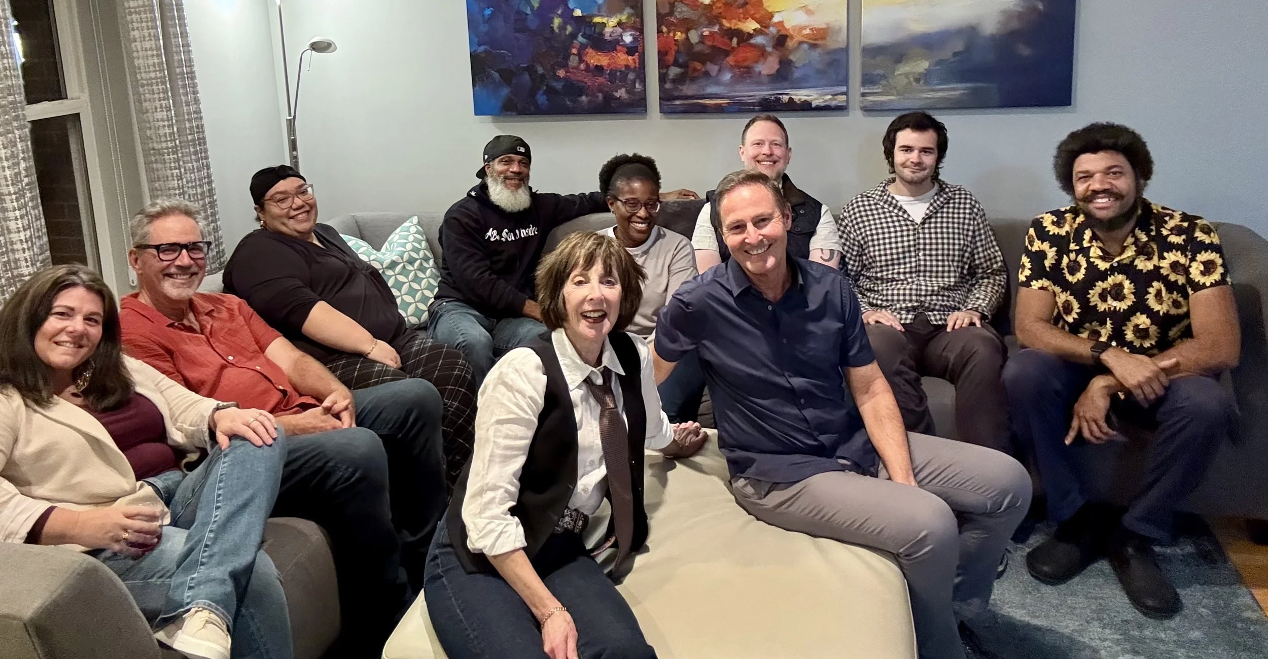 A group of ten diverse people, including men and women, sitting and kneeling on a couch and chairs in a living room, smiling and posing for a photo. The room has a light blue wall, abstract art hanging, and patterned curtains.