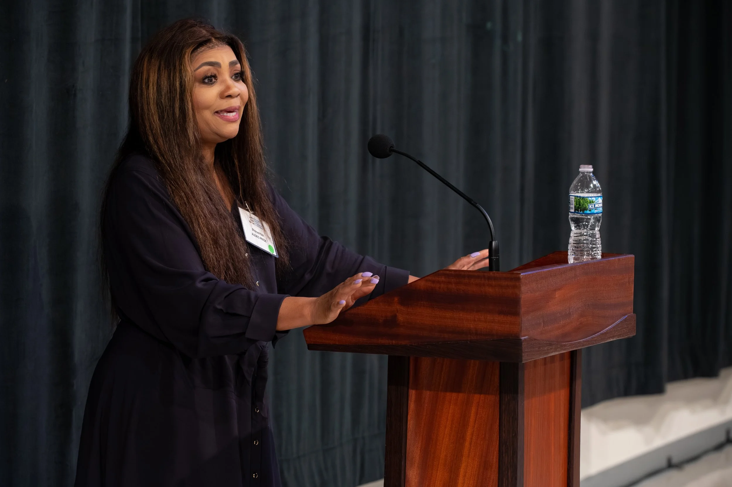 A woman speaking at a podium with a microphone and a water bottle, with a dark curtain background.