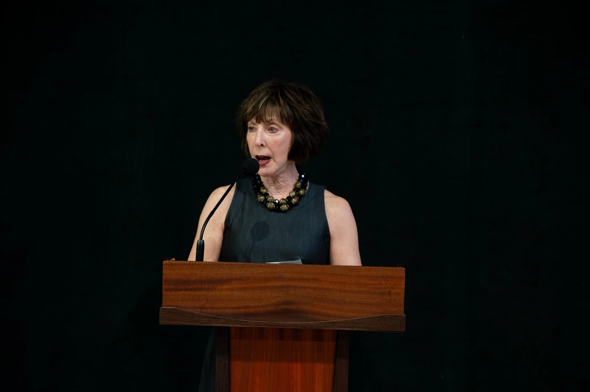 A woman with short brown hair and a black sleeveless dress standing at a wooden podium, speaking into a microphone against a black background.
