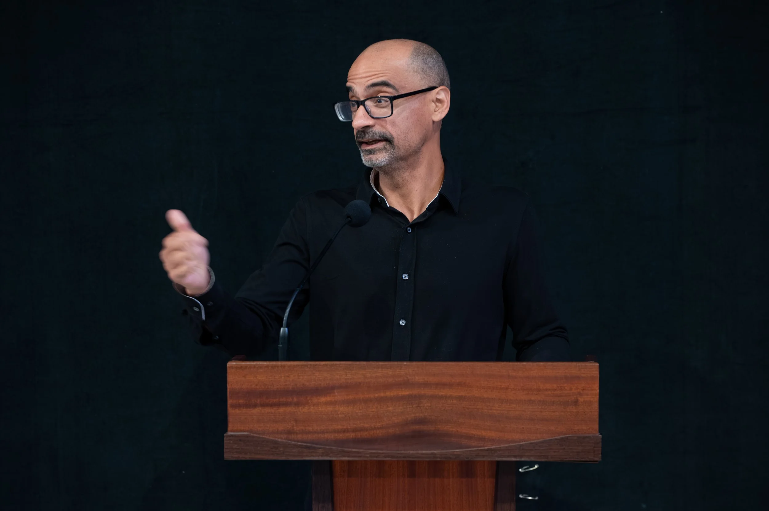A man with glasses, a shaved head, and a beard speaking at a podium in front of a dark background.