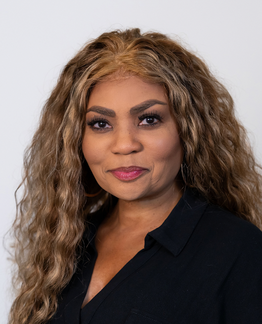 A woman with long, curly, blonde hair and medium skin tone wearing a black collared shirt, standing against a plain white background.