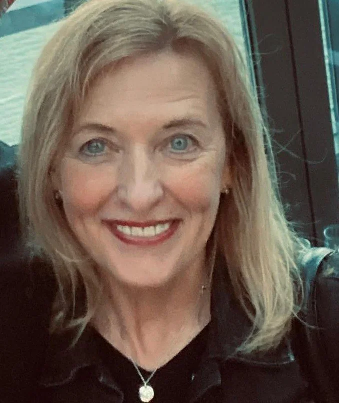 A smiling woman with blonde hair and blue eyes, wearing a black top and silver necklace, in an indoor setting near a window.