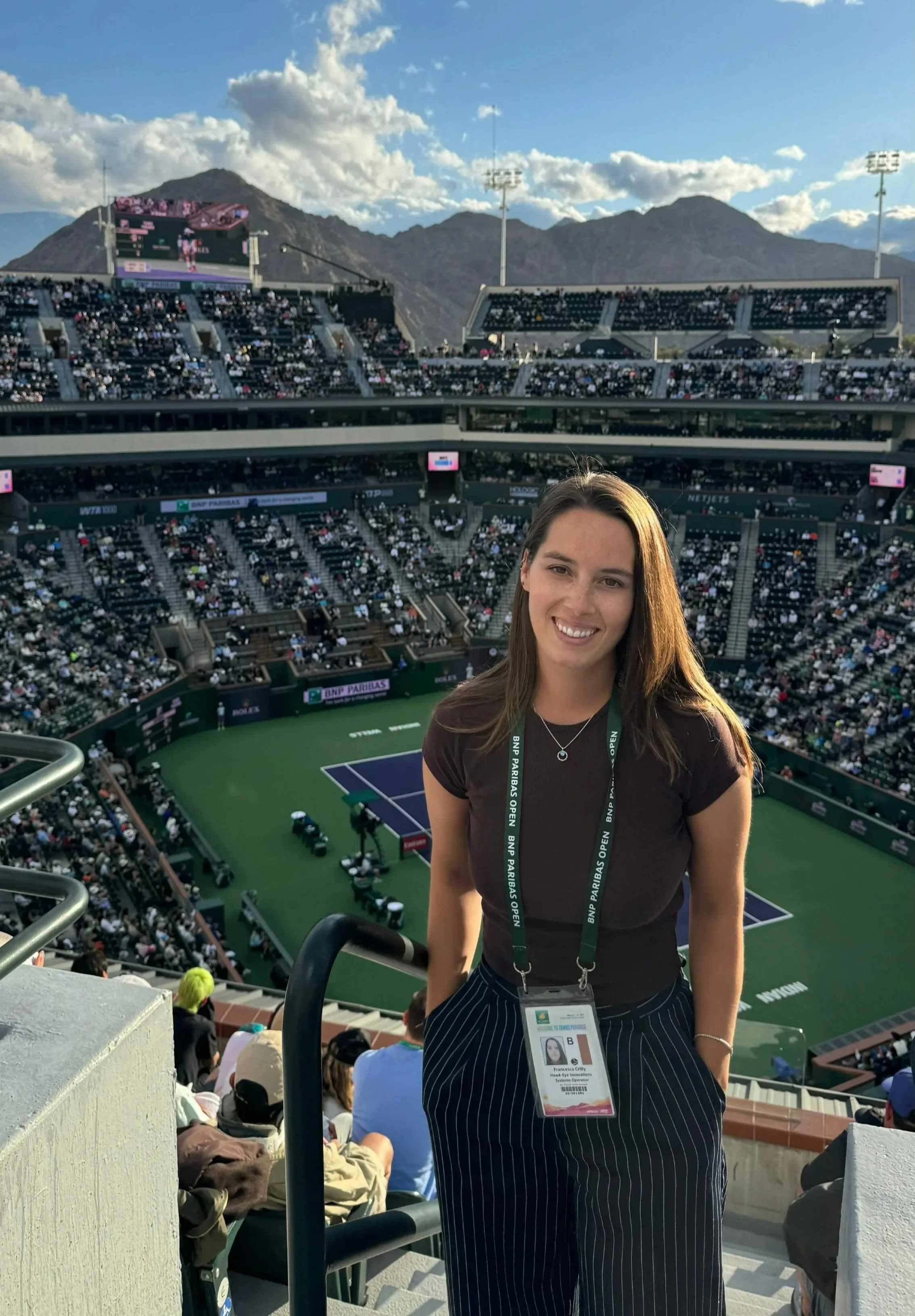 A young woman with brown hair, smiling, wearing a black t-shirt and striped pants, standing in a stadium with a tennis court in the background during a match, with mountains and a partly cloudy sky behind her.