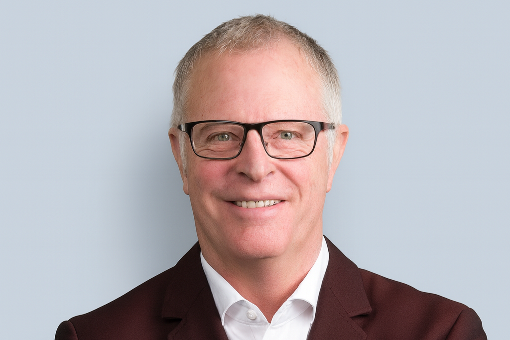 Portrait of a middle-aged man with glasses, wearing a dark suit and white shirt, smiling against a plain light blue background.