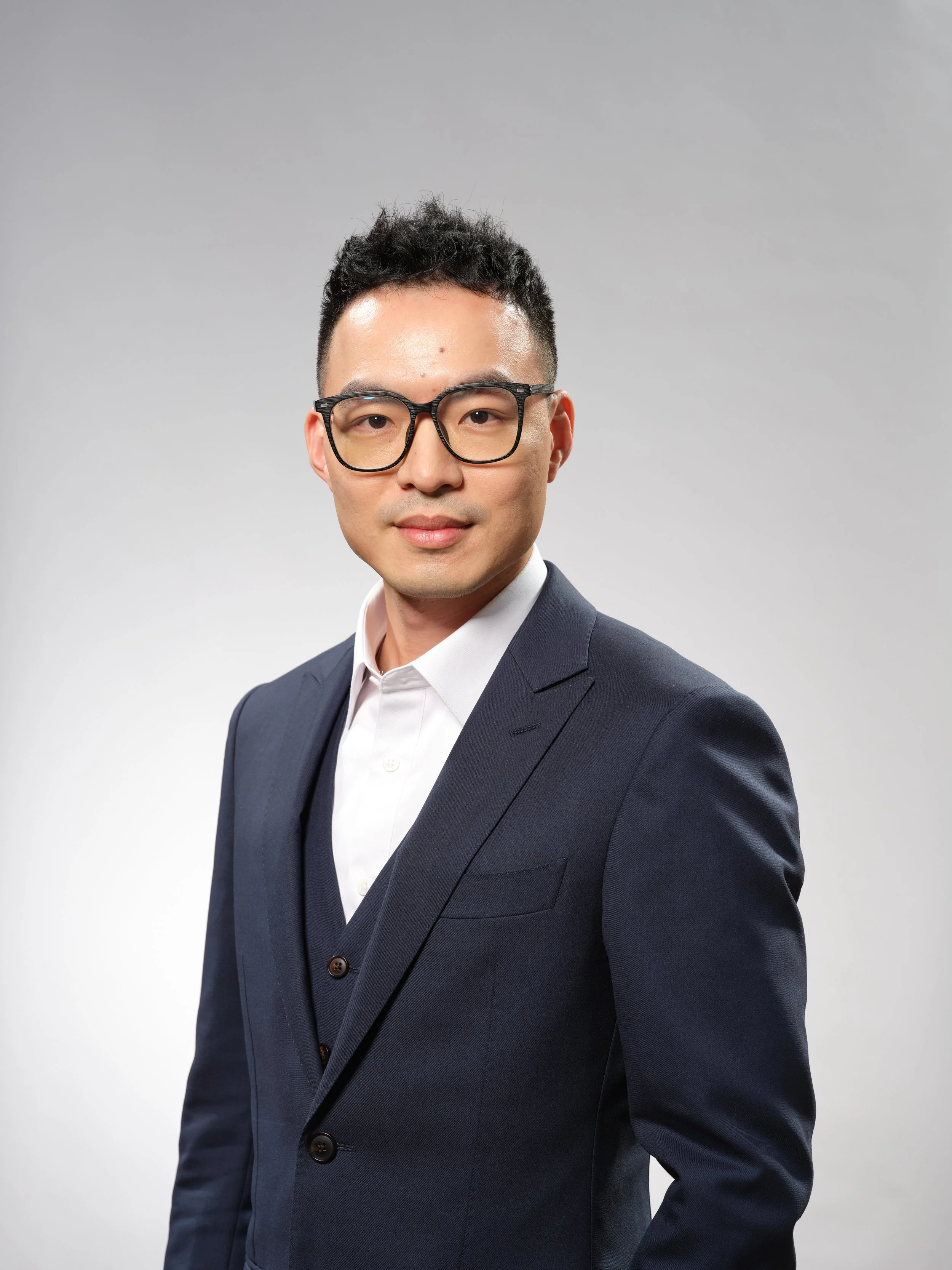 Portrait of a young man with short black hair and glasses, wearing a grey shirt, against a dark background.