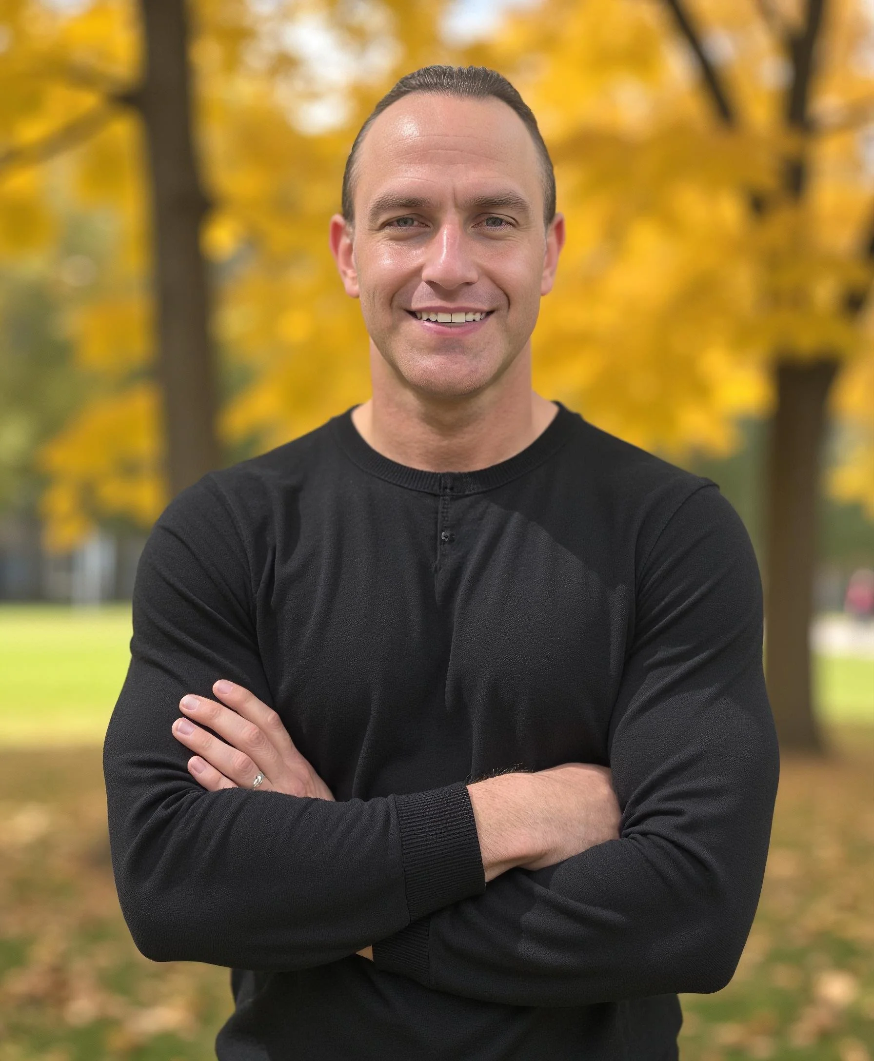 A smiling man with arms crossed, wearing a black long-sleeve shirt, standing outdoors in front of trees with yellow autumn leaves.