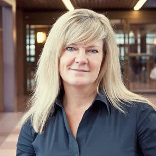 Portrait of a woman with blonde hair, wearing a dark blue blouse, standing indoors in a well-lit space with wooden elements.