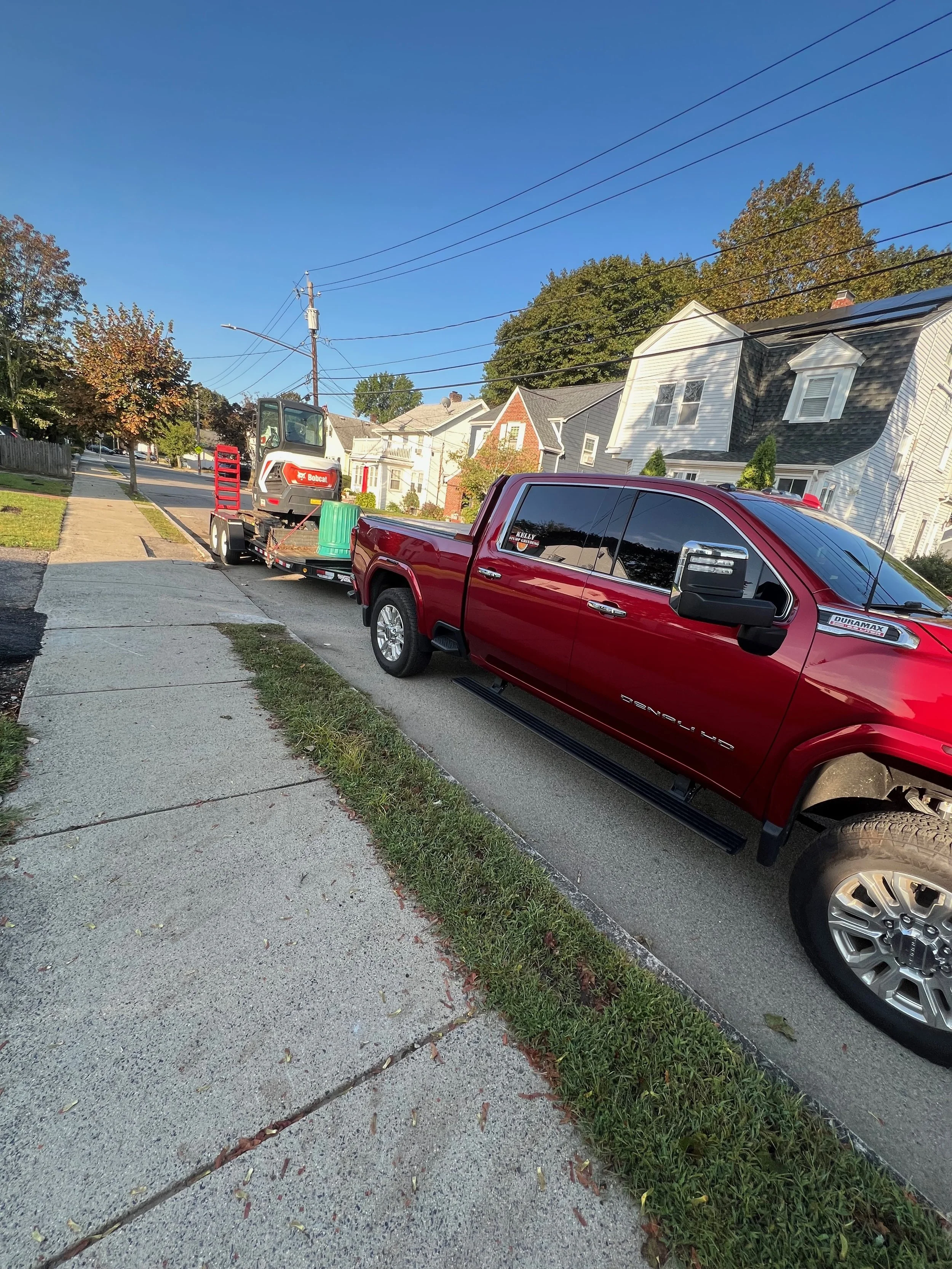 A red pickup truck parked on the side of a residential street, towing a small utility loader on a trailer.
