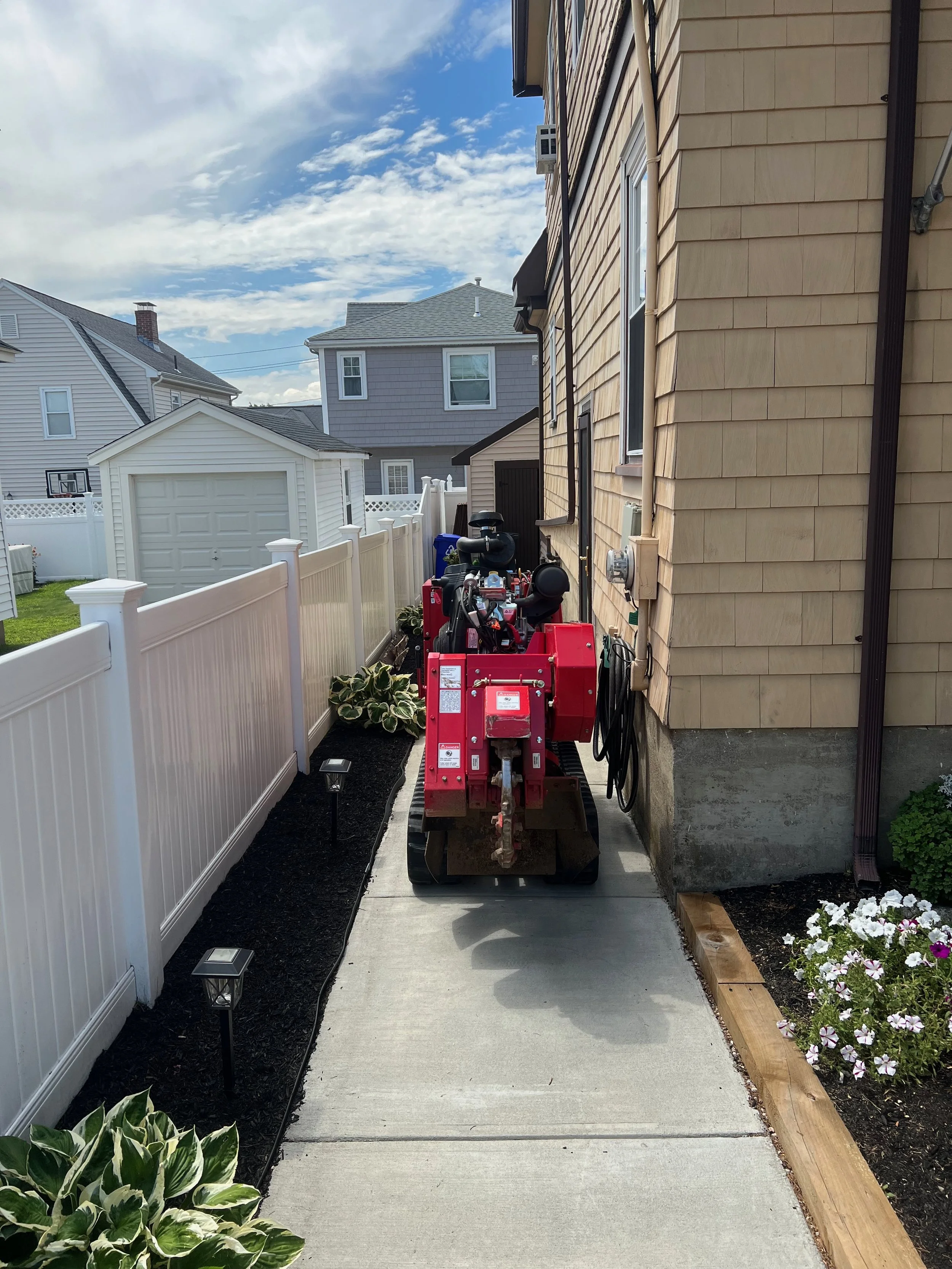 A narrow concrete walkway along the side of a house with a red skid-steer loader parked on it. On the left, there's a white vinyl fence with landscaped plants and small pathway lights. The house has beige siding, and neighboring houses are visible in