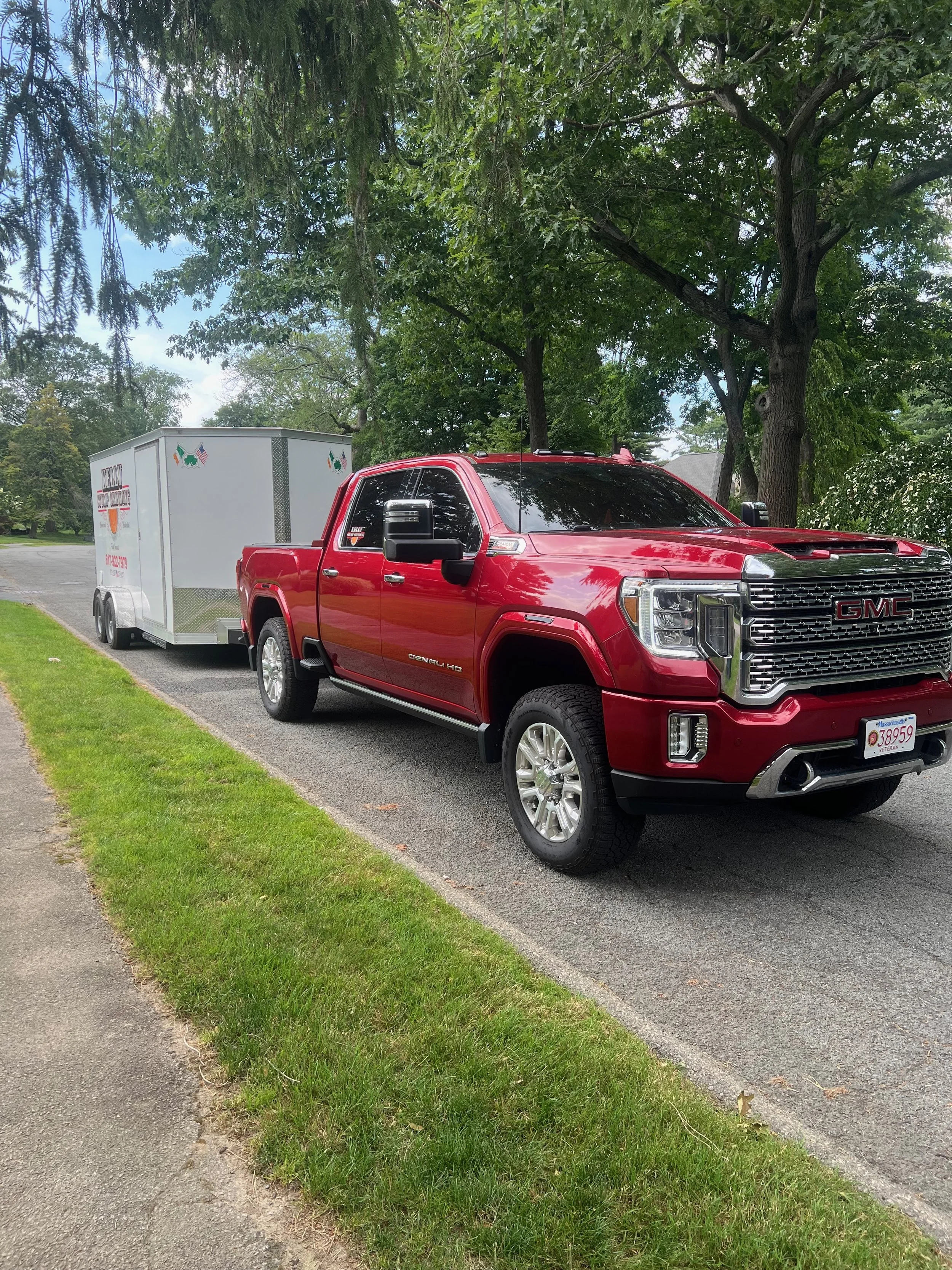 Red GMC pickup truck towing a white enclosed trailer parked on a residential street next to a grassy area with trees in the background.