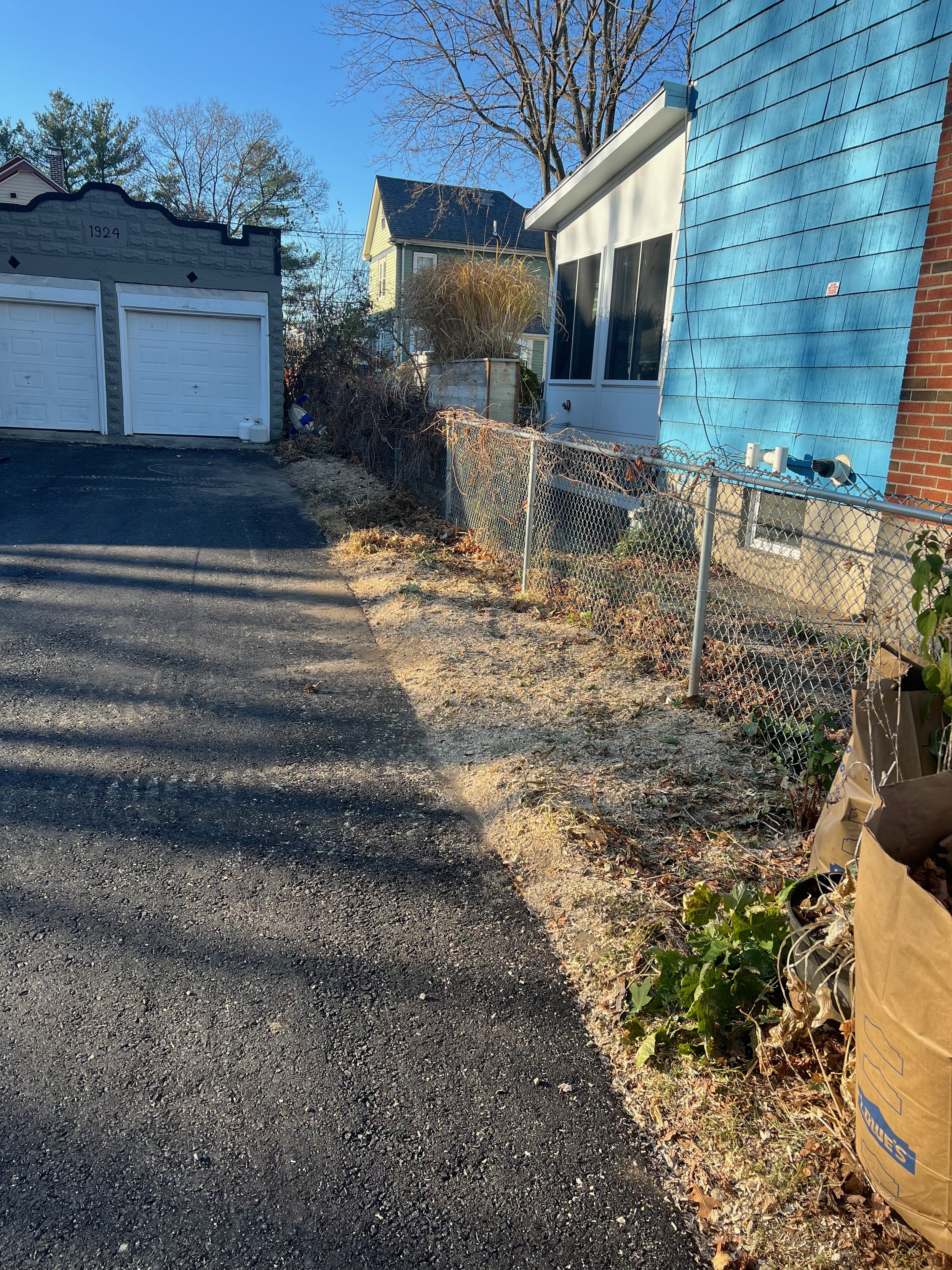 A small driveway in a residential neighborhood with a black asphalt surface, leading to a white garage door. On the right side, there's a chain-link fence, overgrown with dry plants and leaf debris along the edge of the driveway. Behind the fence, a 