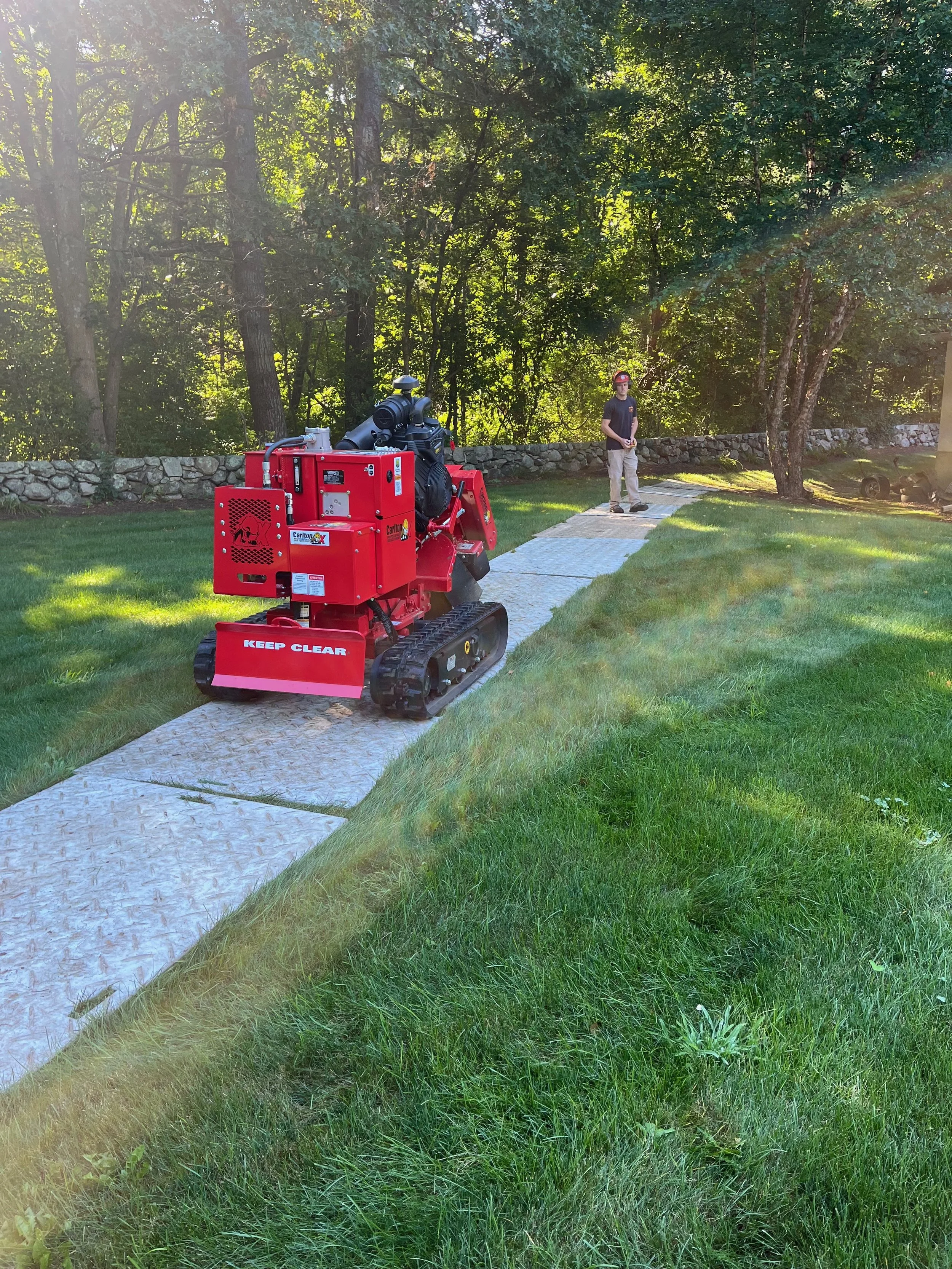 A person watching a small red tracked robot on a concrete pathway in a park-like setting with green grass, trees, and a stone wall in the background.
